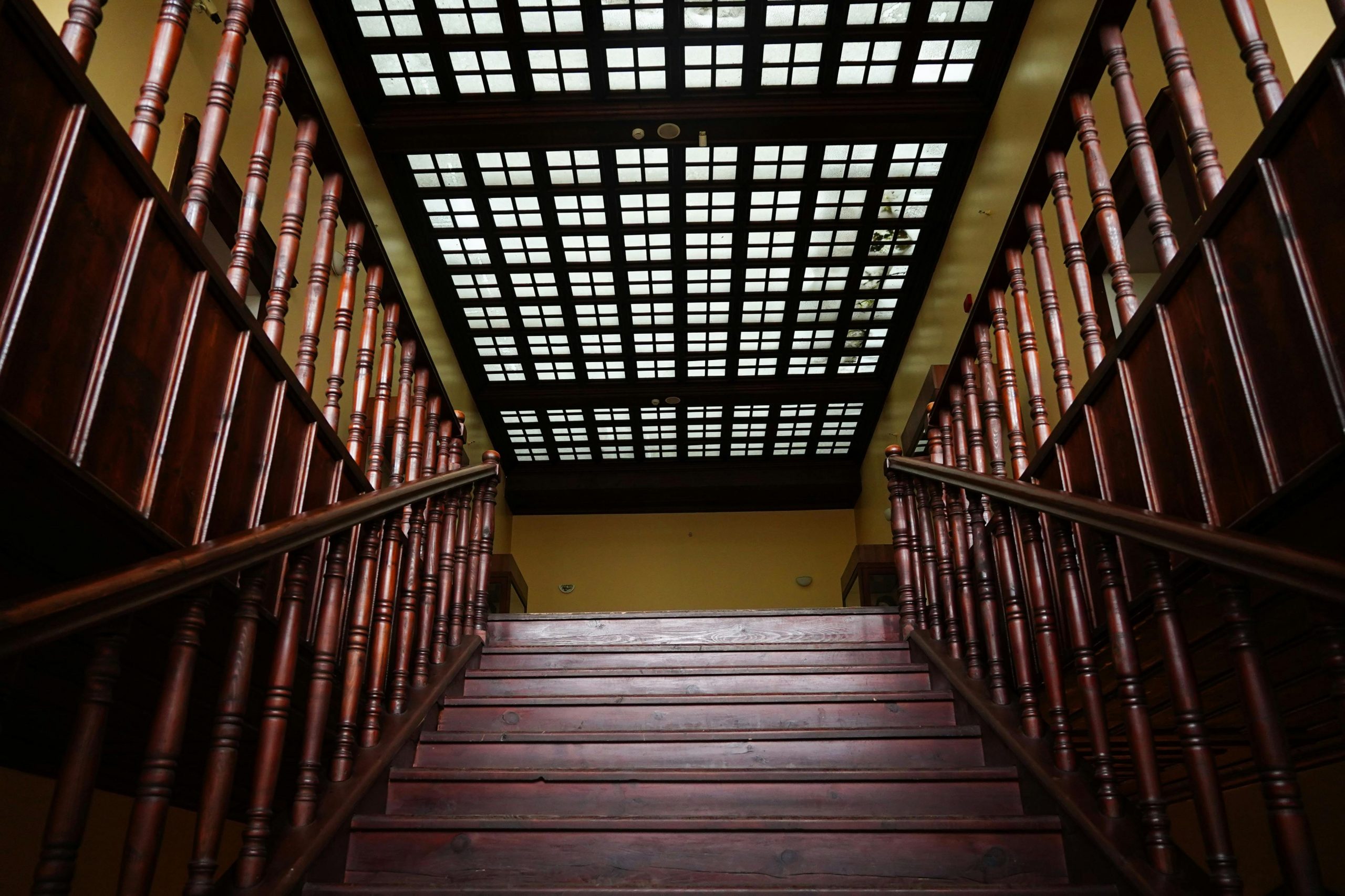 A grand wooden staircase ascends to a grid-patterned glass ceiling in an elegant interior.