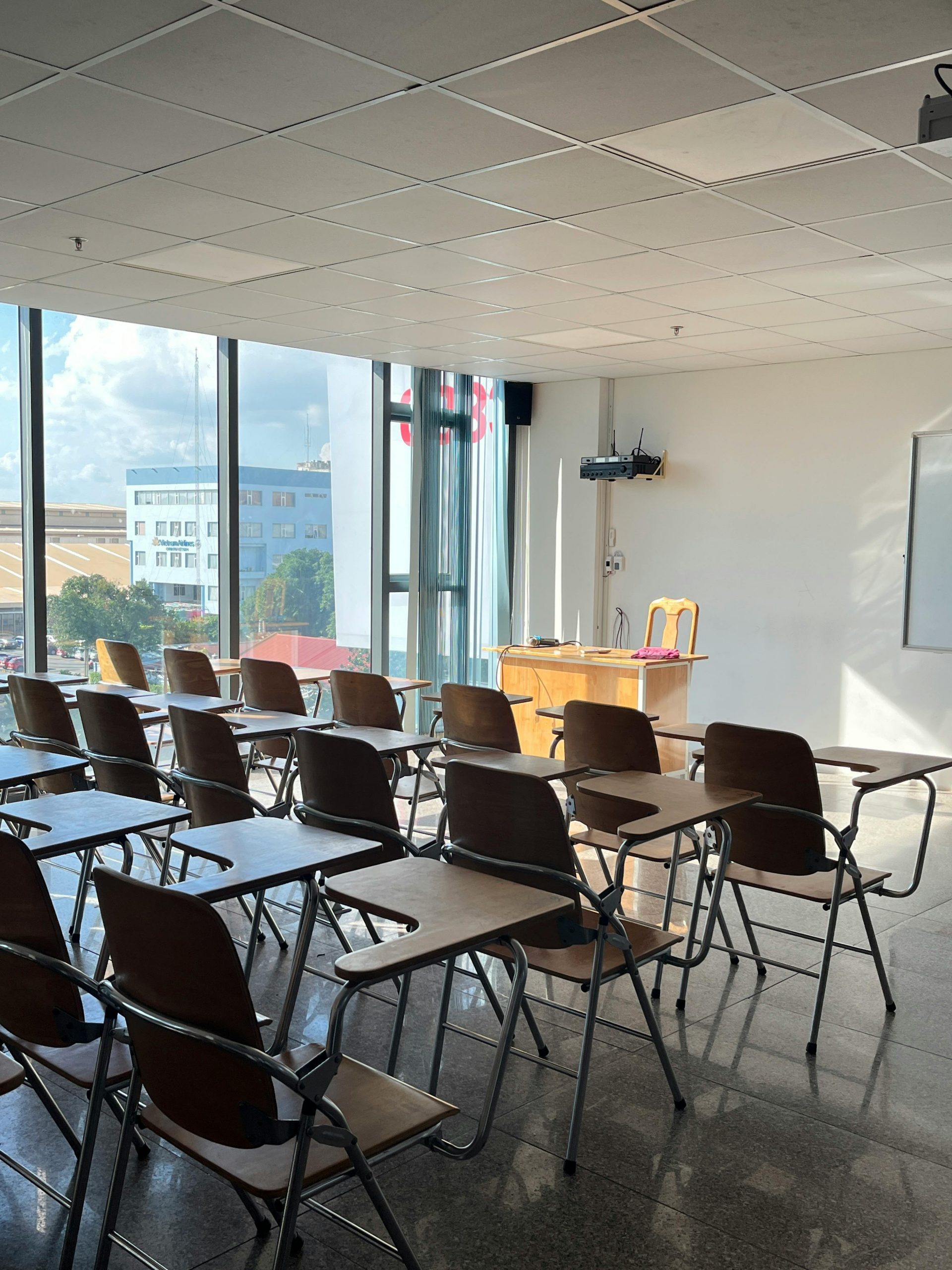 Bright and empty classroom with sunlight streaming through large windows, featuring wooden desks and chairs.