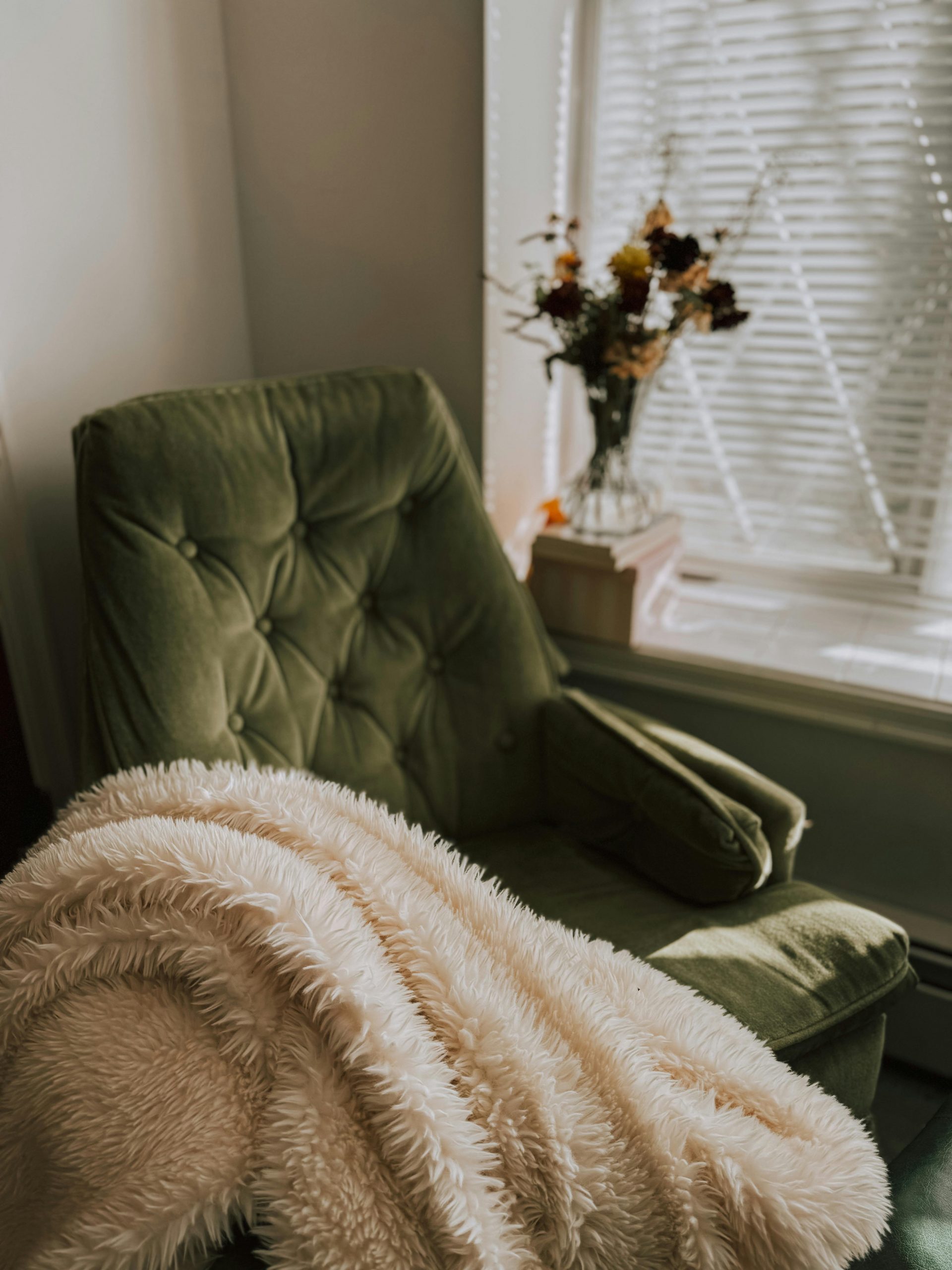 Green armchair with a fluffy blanket and flowers by the window, bathed in soft sunlight.