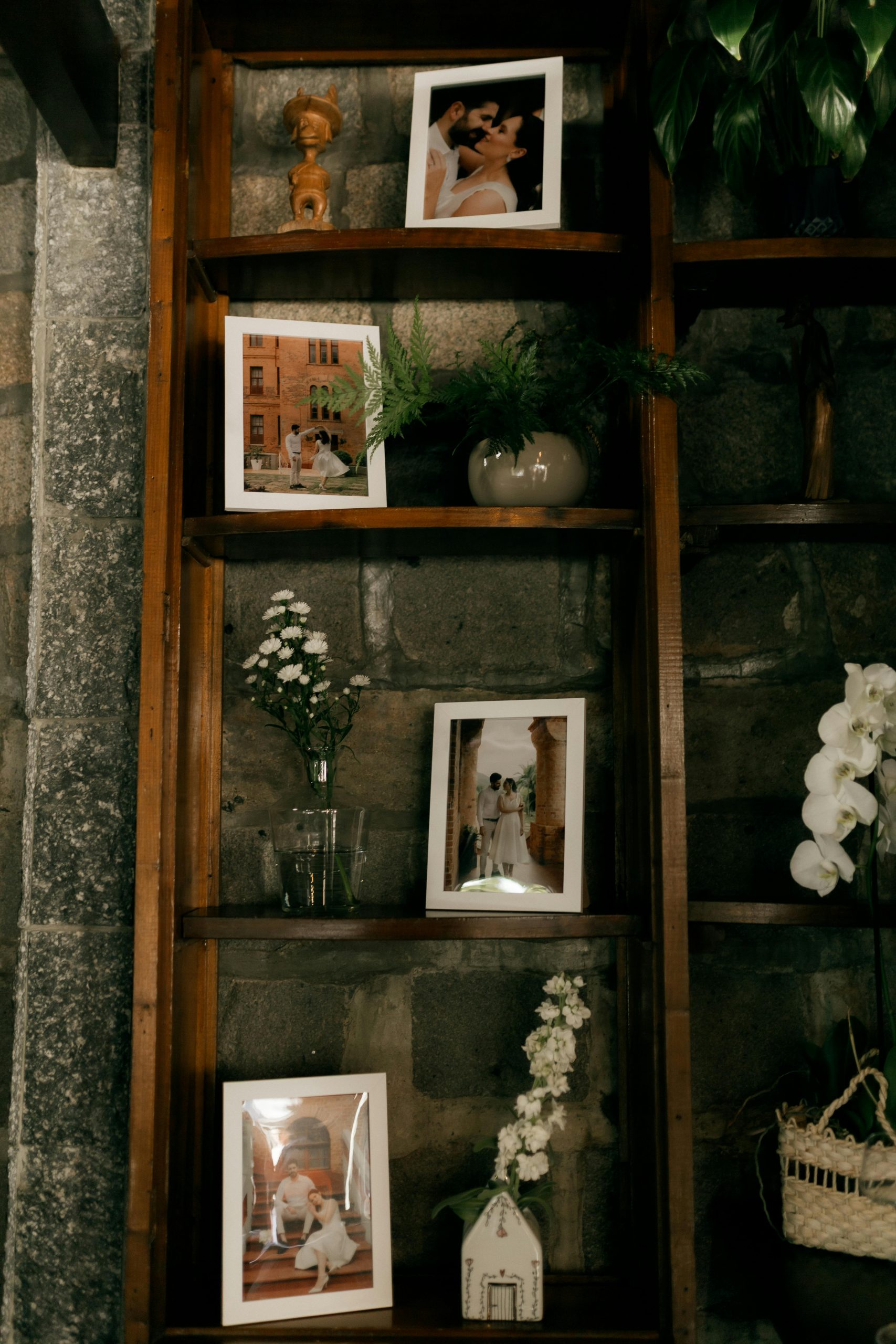 Wooden shelf featuring framed photos, plants, and flowers on a stone wall.