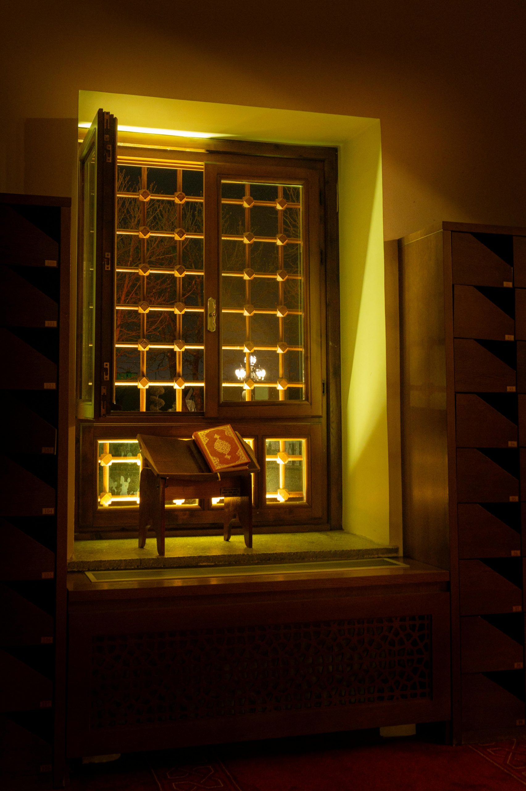 A beautifully lit mosque interior featuring an open Quran on a wooden stand by a window with intricate designs.