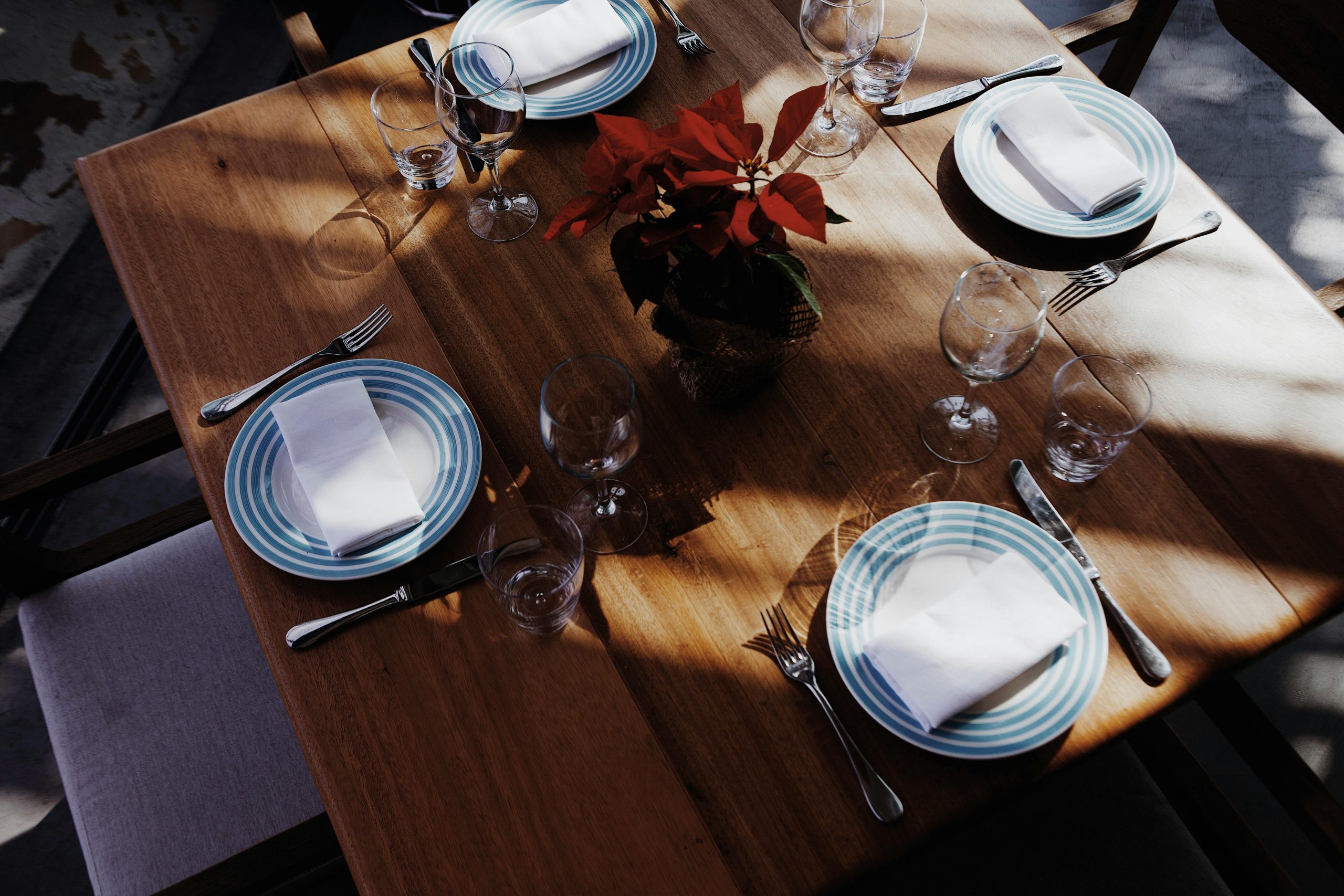 A beautifully set dining table with striped plates and silverware, centered around a red flower arrangement.