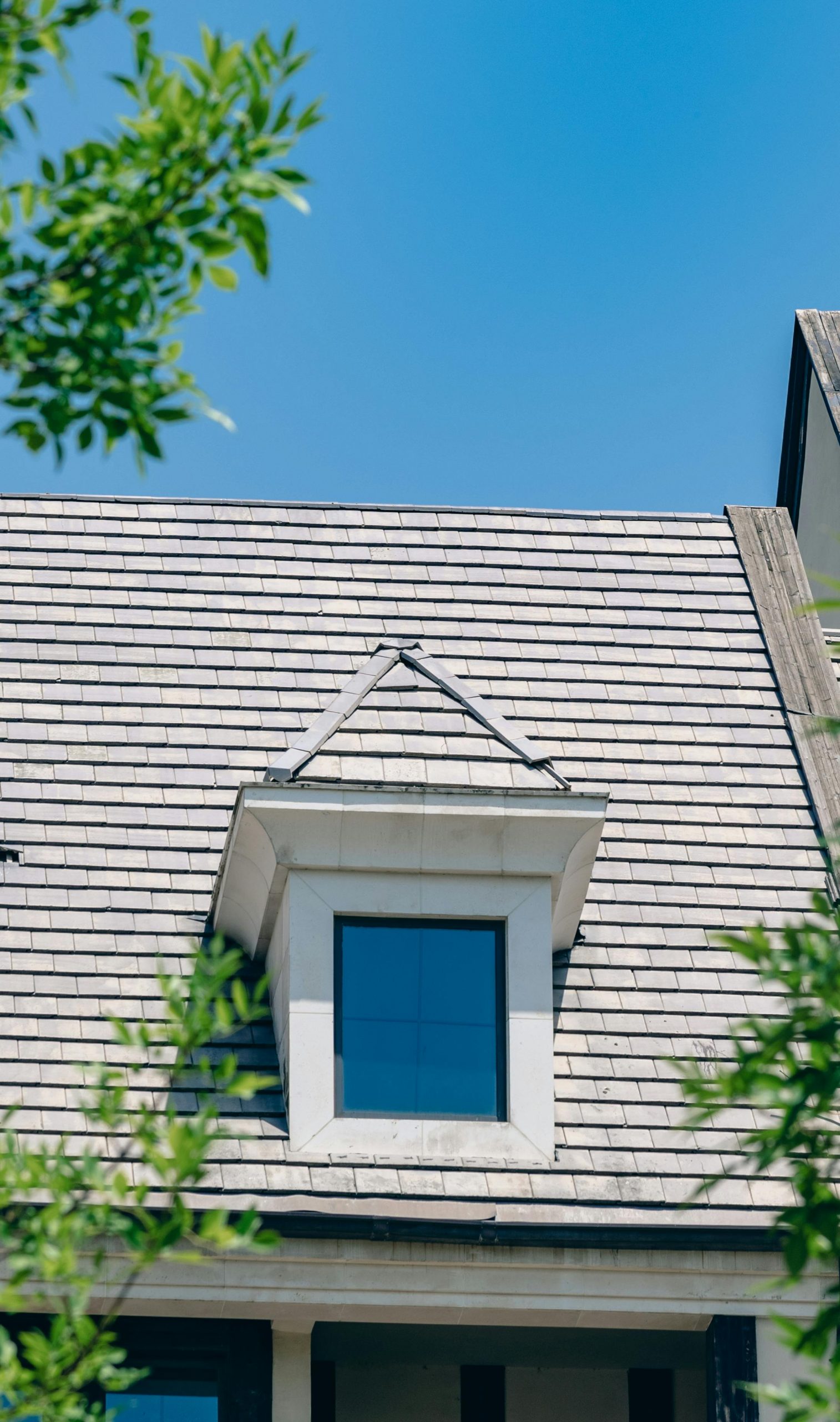 A Cape Cod style house roof with a prominent dormer window under a clear blue sky.