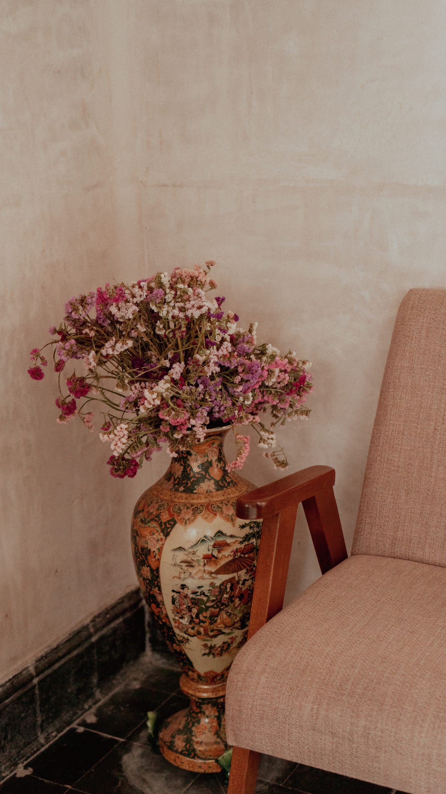 A decorative vase filled with pink flowers beside a beige chair in a minimalist room setting.