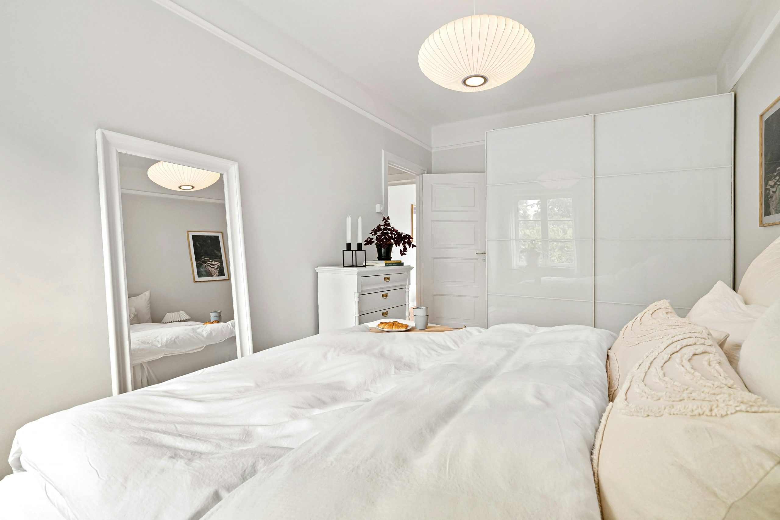 A minimalist bedroom with white decor, featuring a bed, dresser, and mirror under soft lighting.