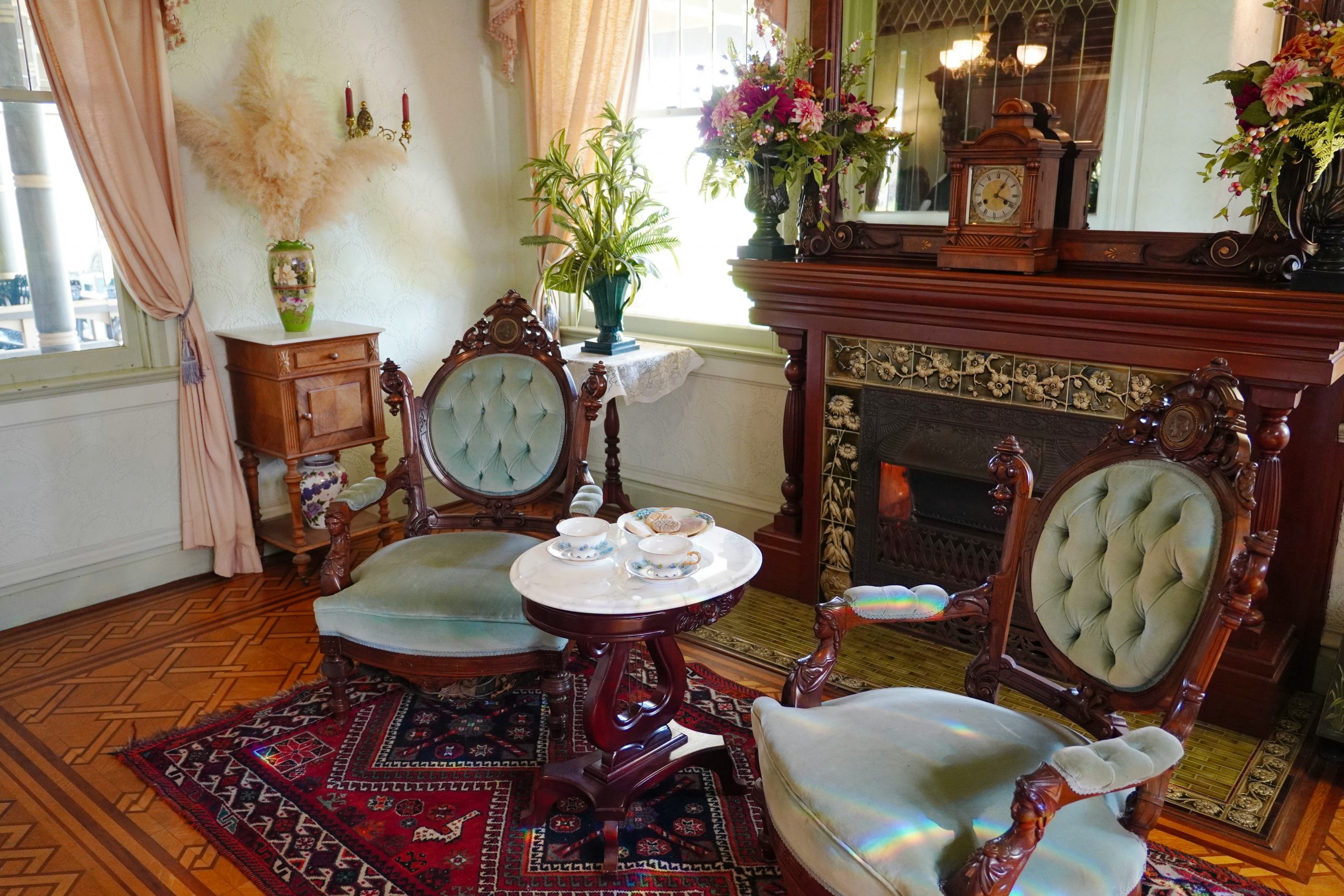 An inviting Victorian parlor at the Winchester Mystery House features carved wood armchairs with tufted velvet upholstery, a marble-topped table set for tea, and a richly tiled fireplace beneath a gra