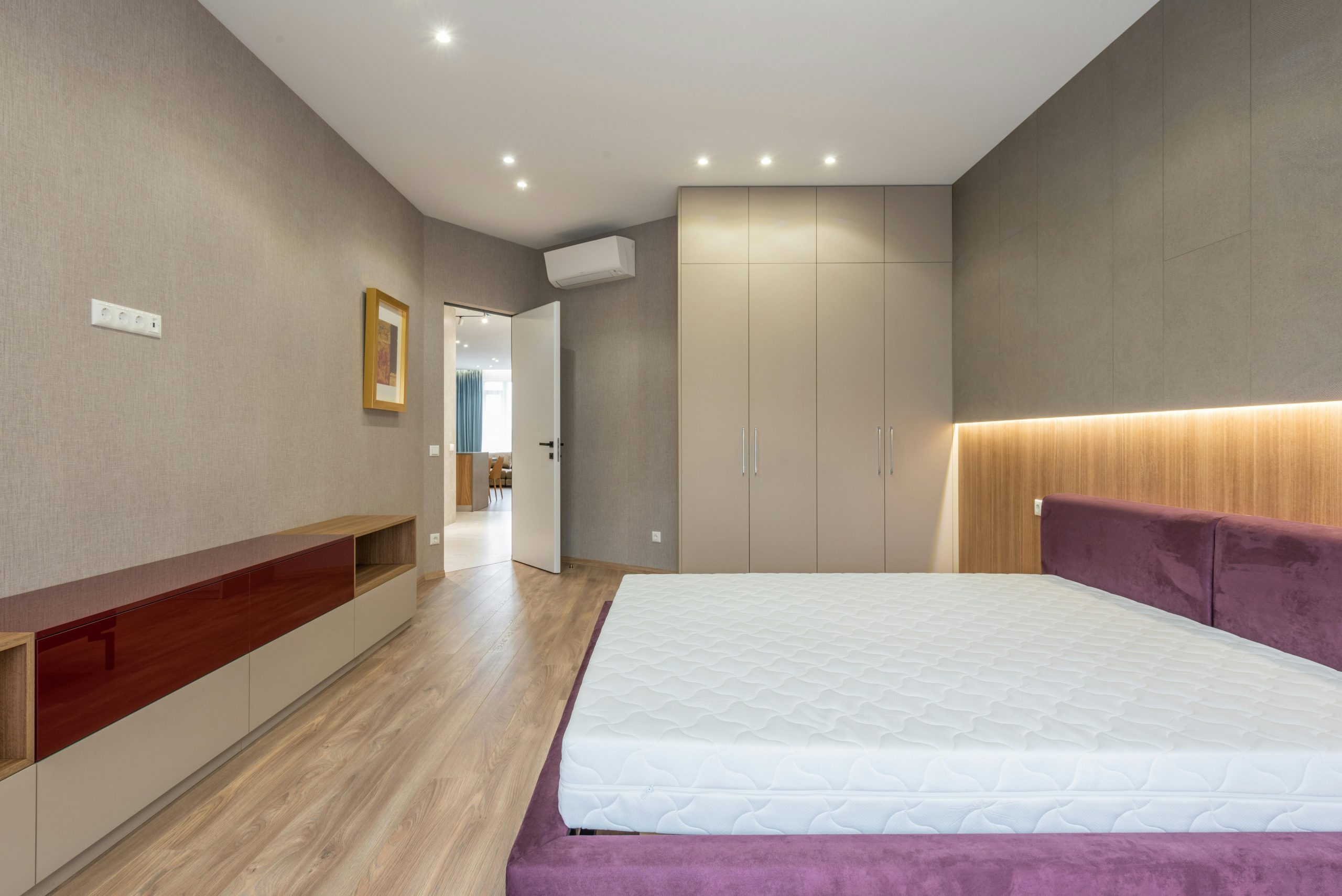 Interior of stylish spacious bedroom with white mattress on bed placed against cupboard near wardrobe and opened door in apartment