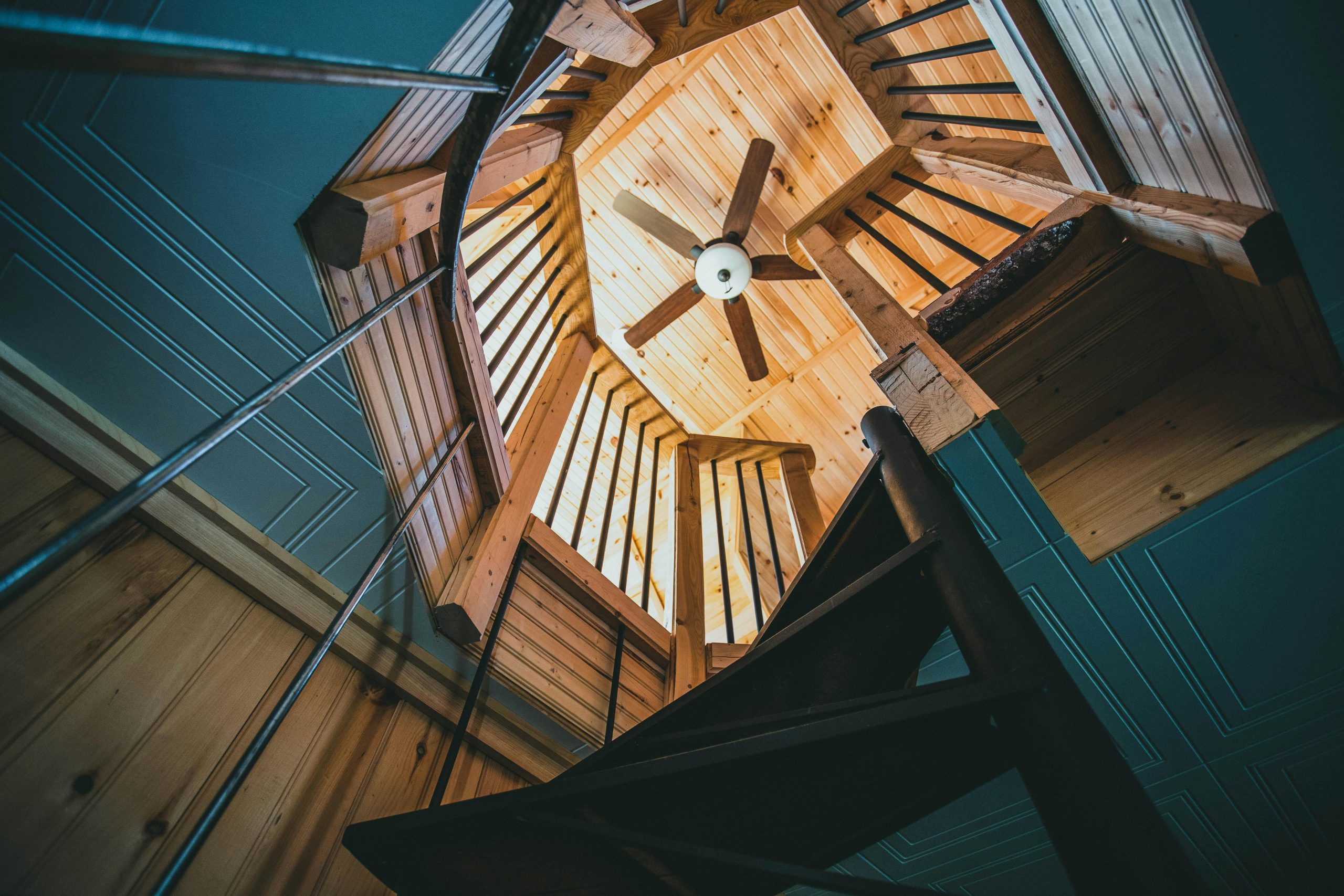Low-angle view of a modern wooden spiral staircase with a ceiling fan in a house interior.