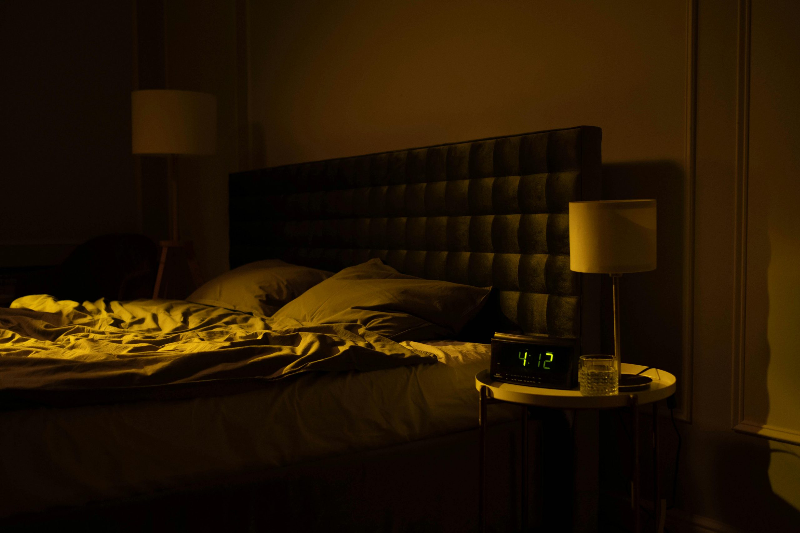 Relaxing dimly lit bedroom with an unmade bed, digital clock displaying 4:12, and warm ambient lighting.