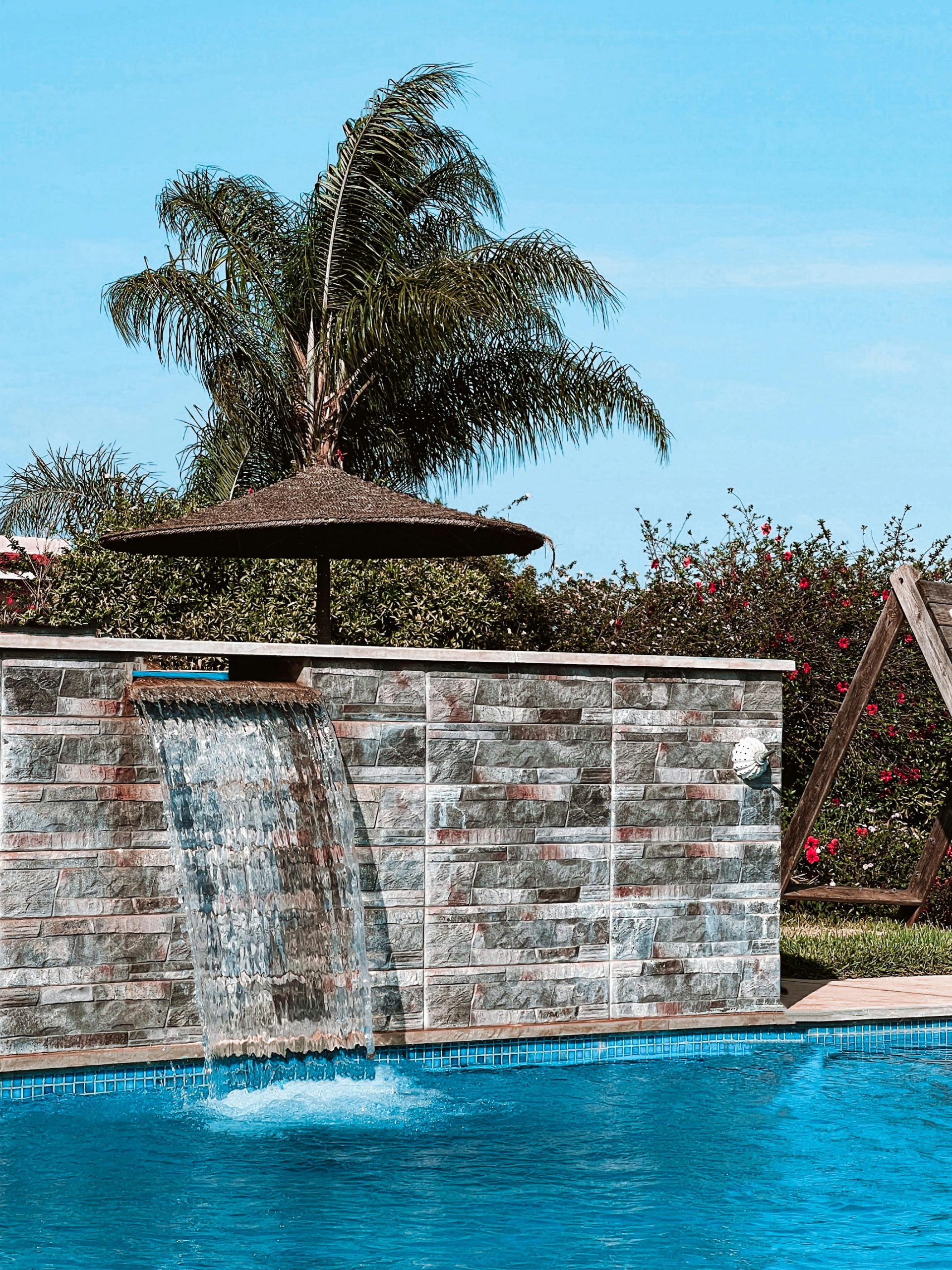 Serene tropical pool area with a flowing waterfall and lush palm trees under clear blue sky.