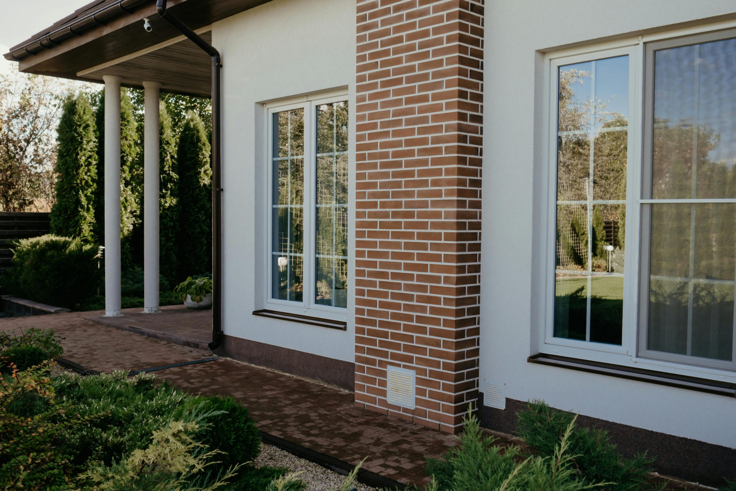 Stylish modern house facade with brick pillar, large glass windows, and lush greenery.
