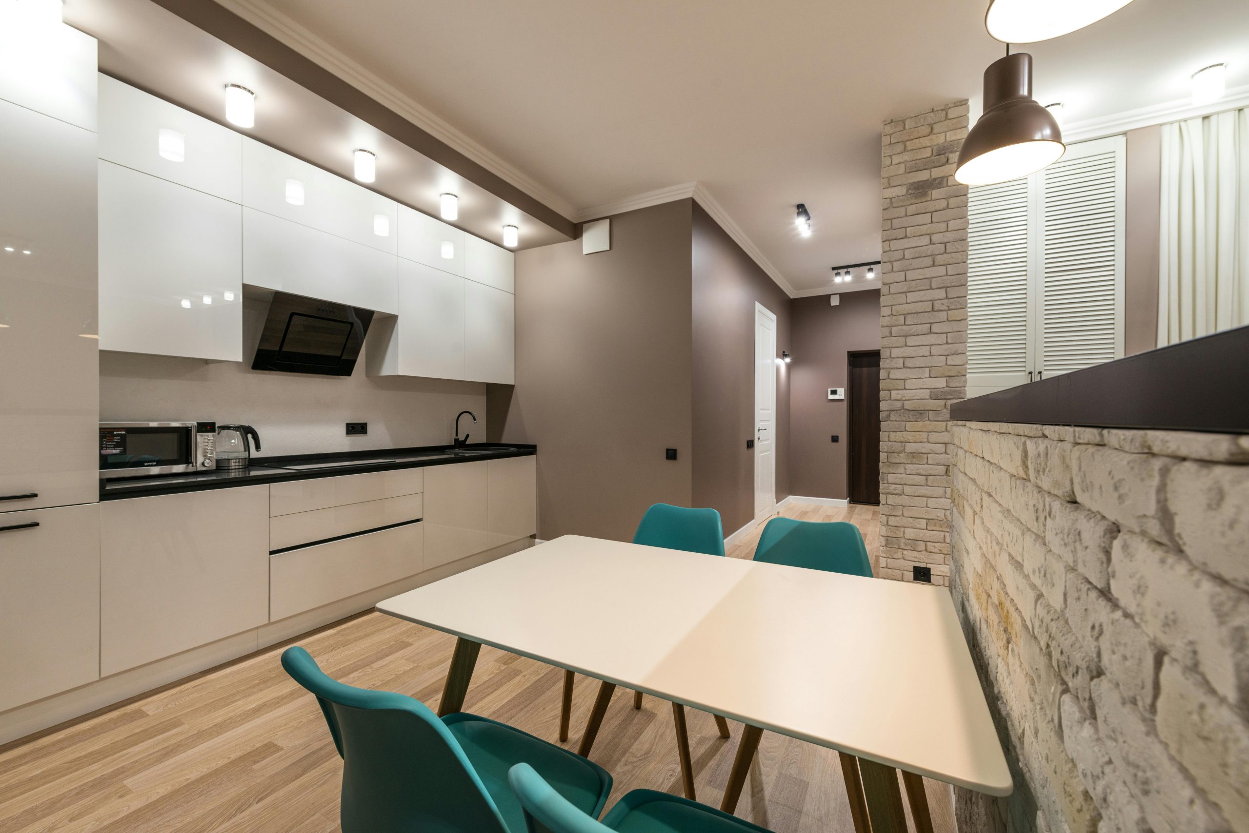 Table with chairs placed in modern kitchen with white cupboards and counter with appliances in spacious apartment with glowing lights