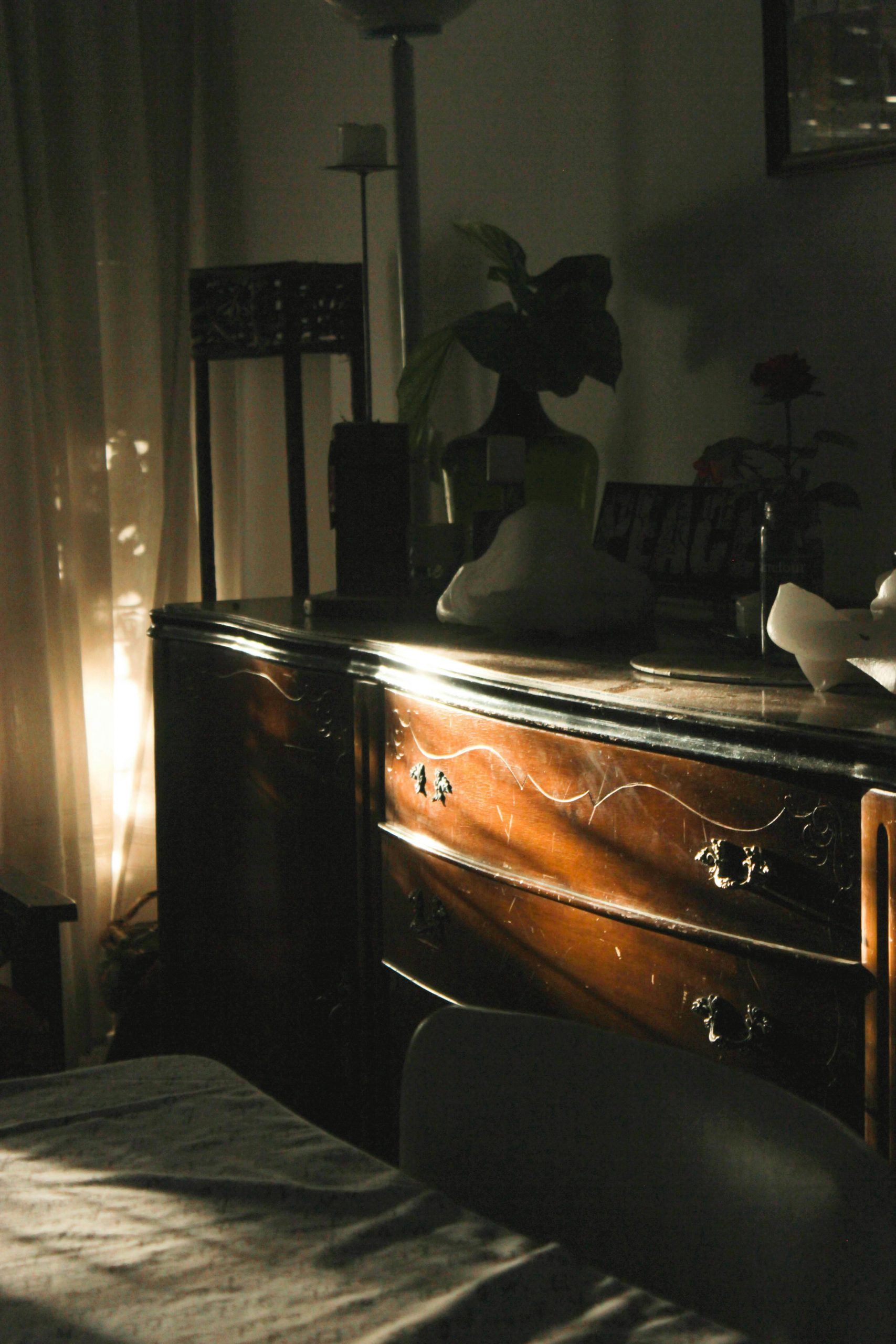 Warm sunlight filters through curtains, casting shadows on a wooden dresser in a cozy interior.