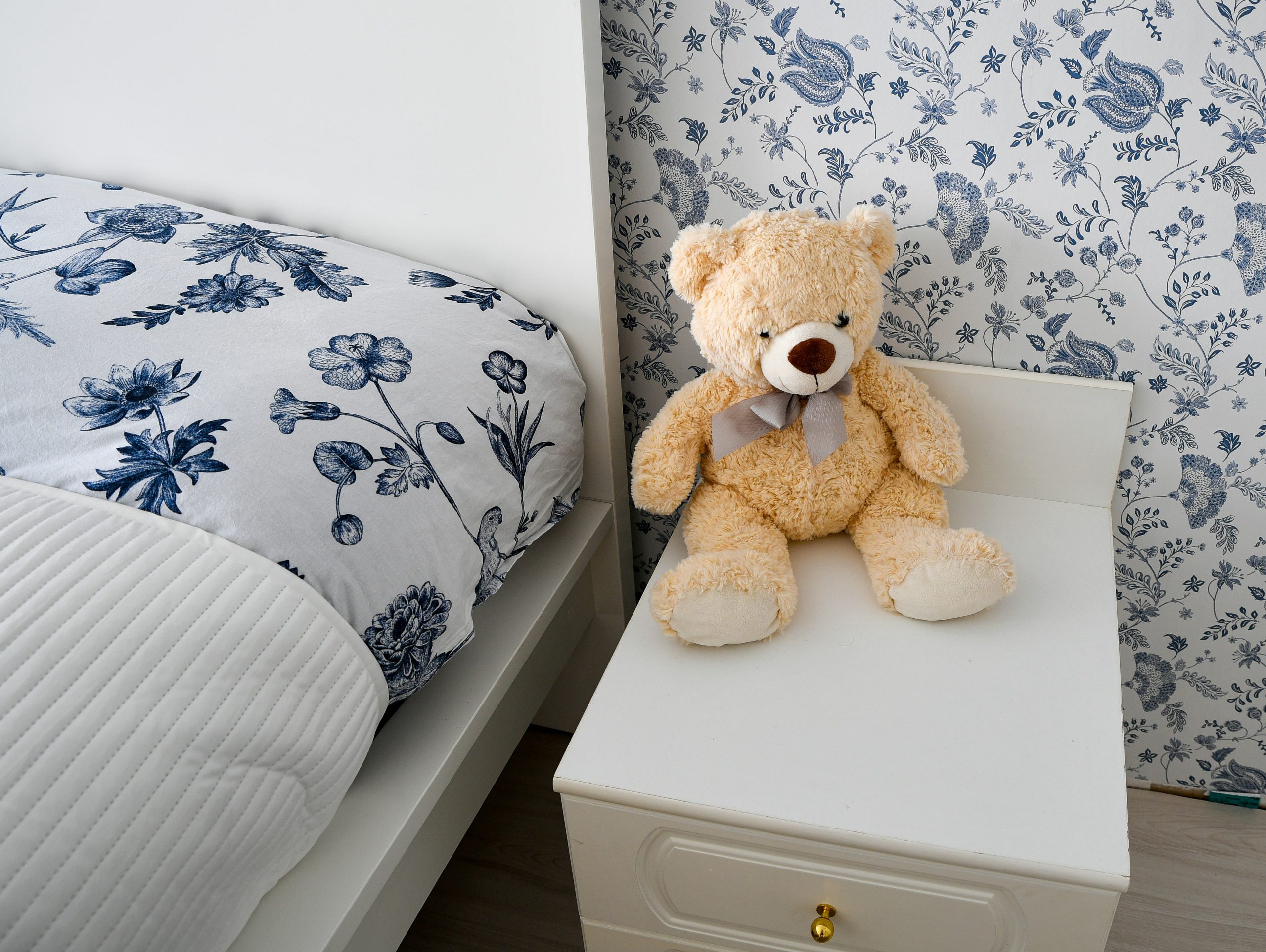 A cute fluffy teddy bear sitting on a white nightstand next to a neatly made bed with blue and white floral patterned bedding and matching wallpaper.📸 Photo by me IG: @margoevardson