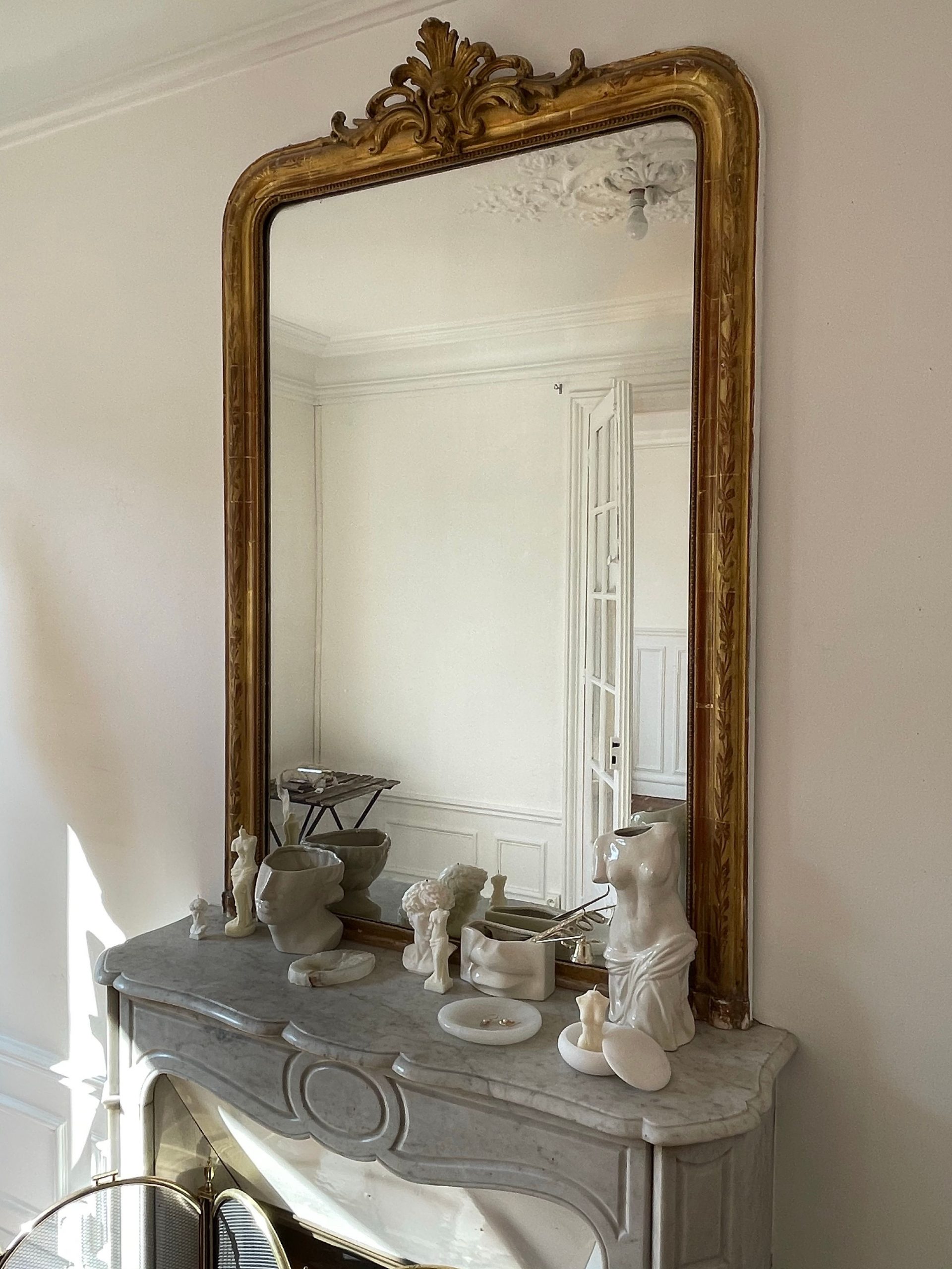 A luxurious room features a large vintage mirror above a marble fireplace with classic decor pieces.
