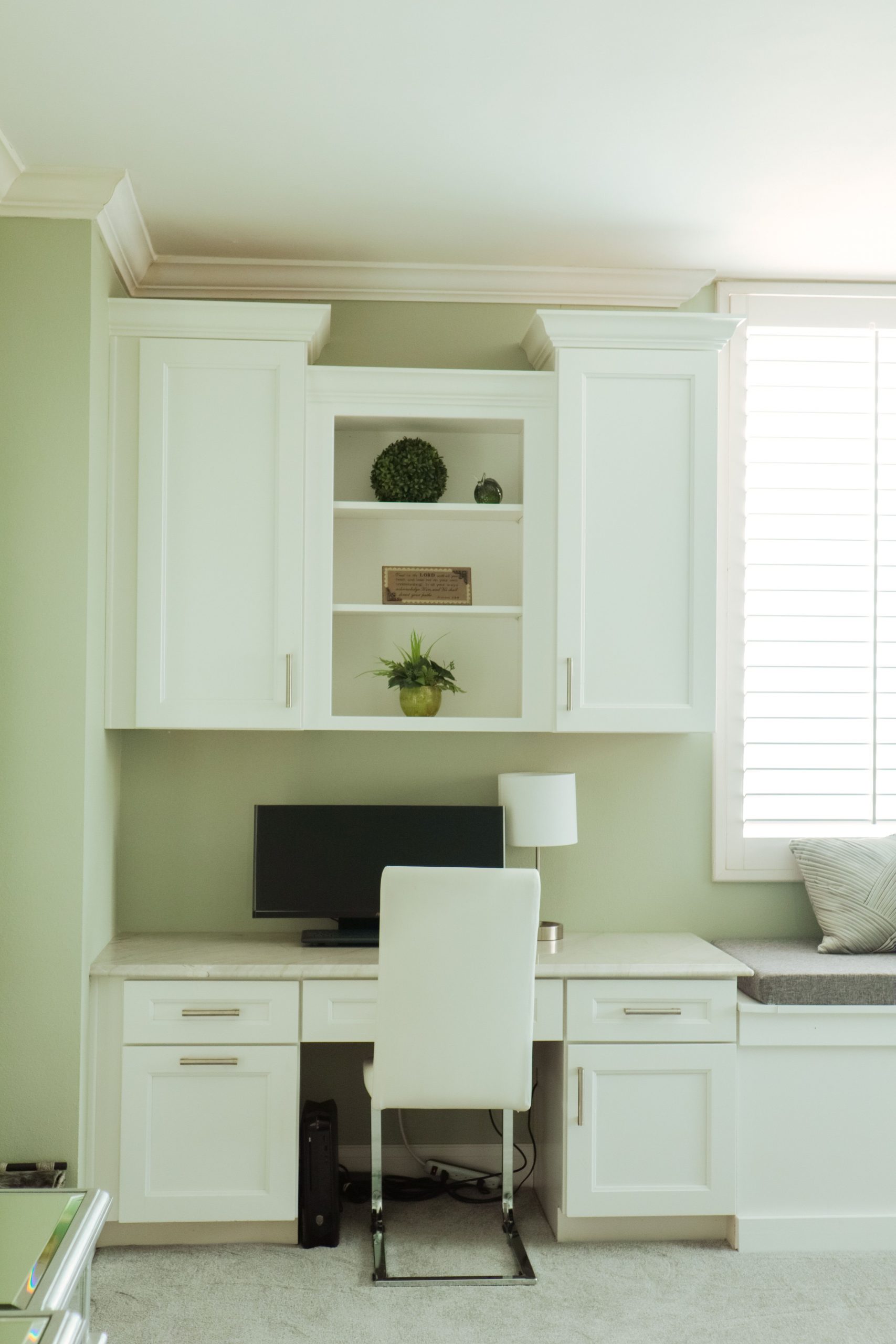 A stylish home workspace with white desk, chair, and cabinets, featuring a cozy window seat.