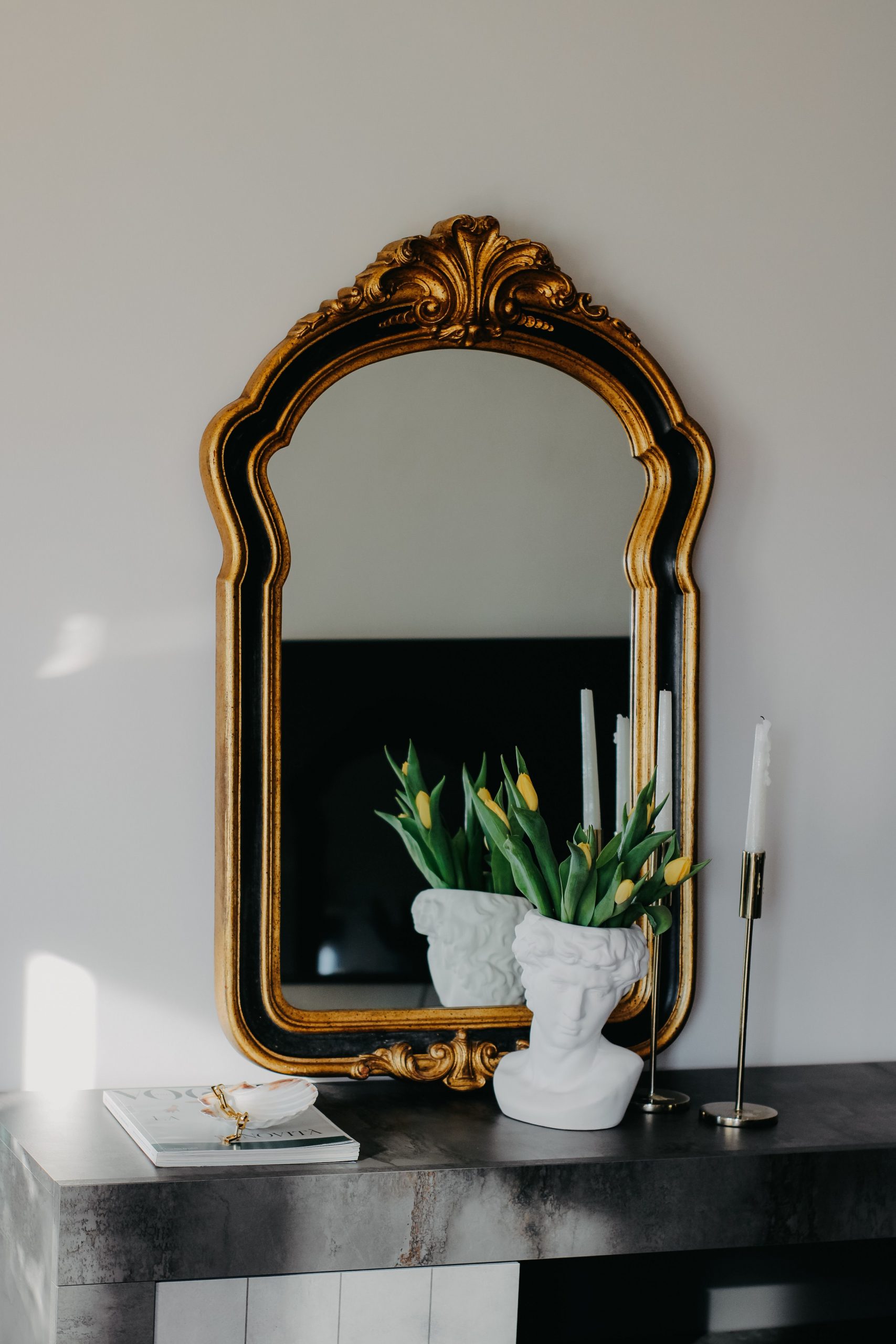 An ornate mirror with flowers, vase, and candles on a modern mantelpiece reflecting elegance.