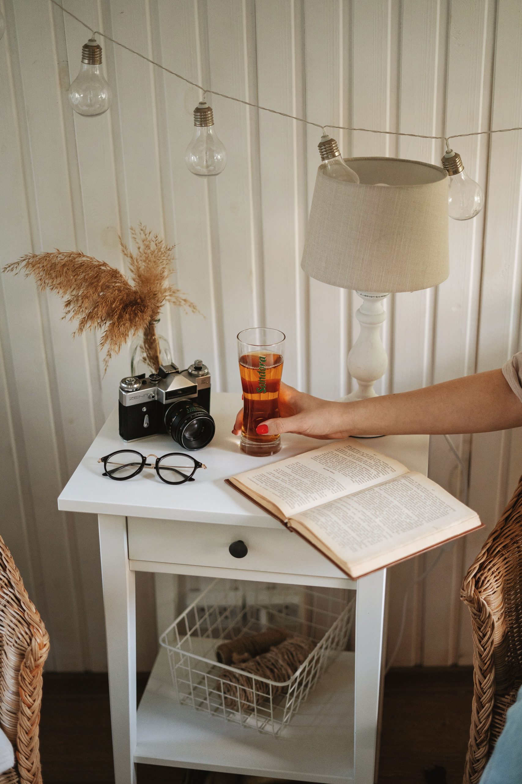 Bedside table with open book, camera, drink, and lamp for a cozy home atmosphere.