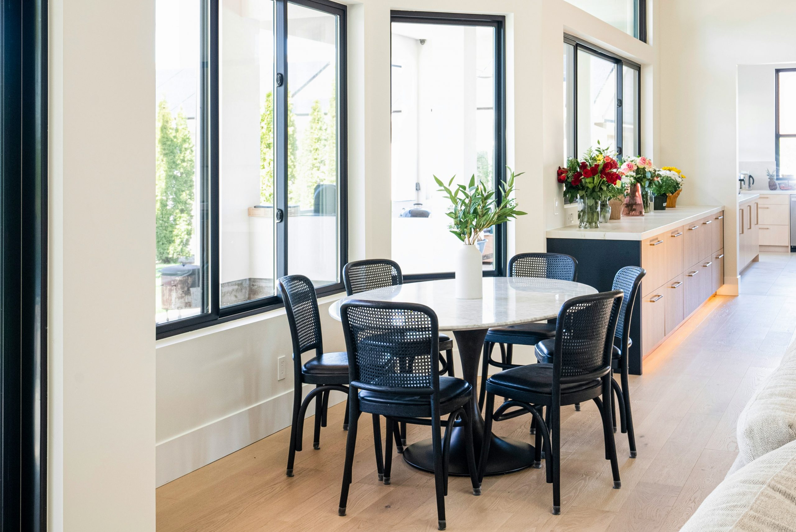 Bright modern dining area with round marble table, black woven chairs, and large floor-to-ceiling windows. Contemporary open-concept home featuring warm wood cabinetry, quartz countertops, and natural