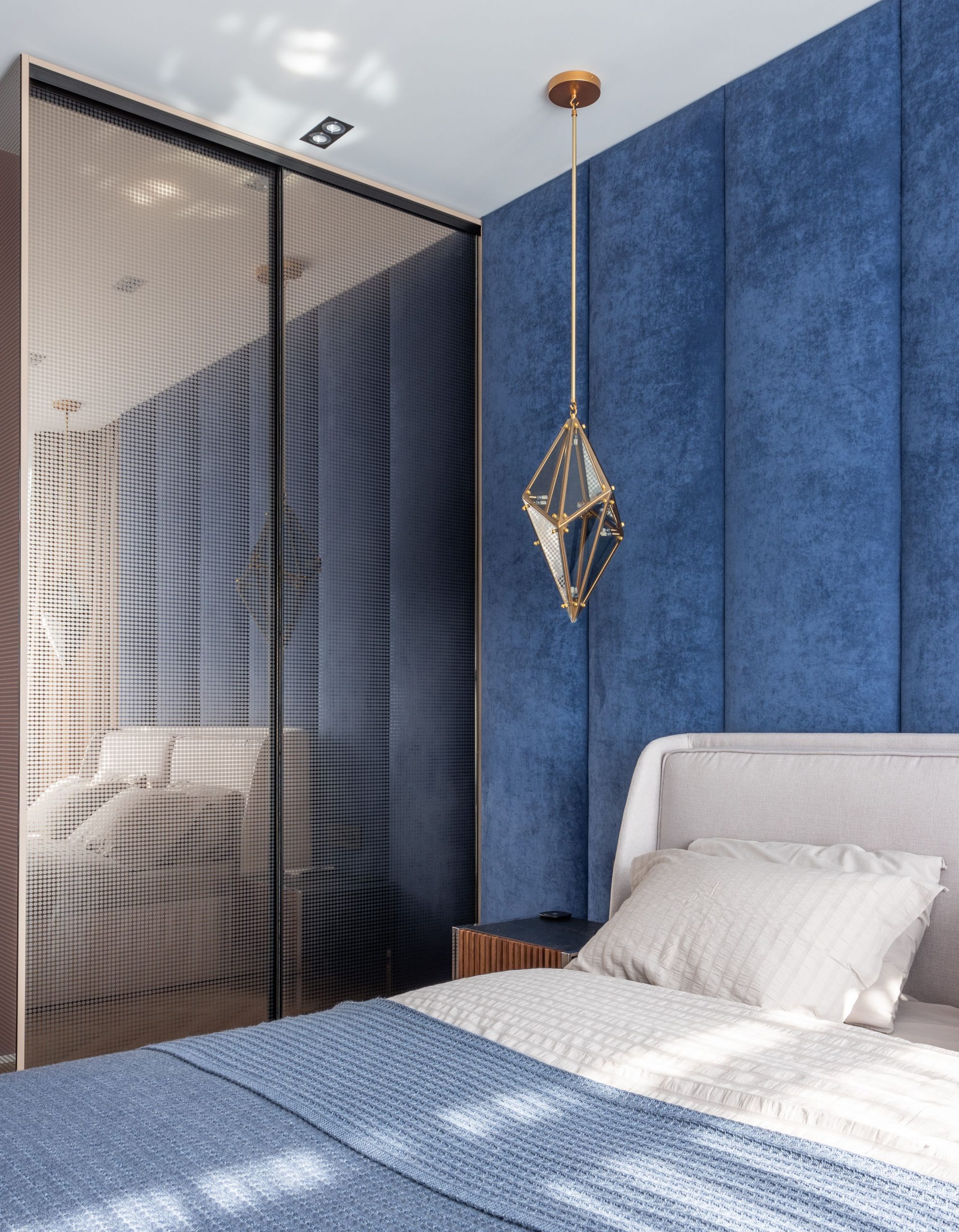 Comfortable bed with cushion and blanket placed at blue wall near big wardrobe in modern bedroom with decorations and sunlight