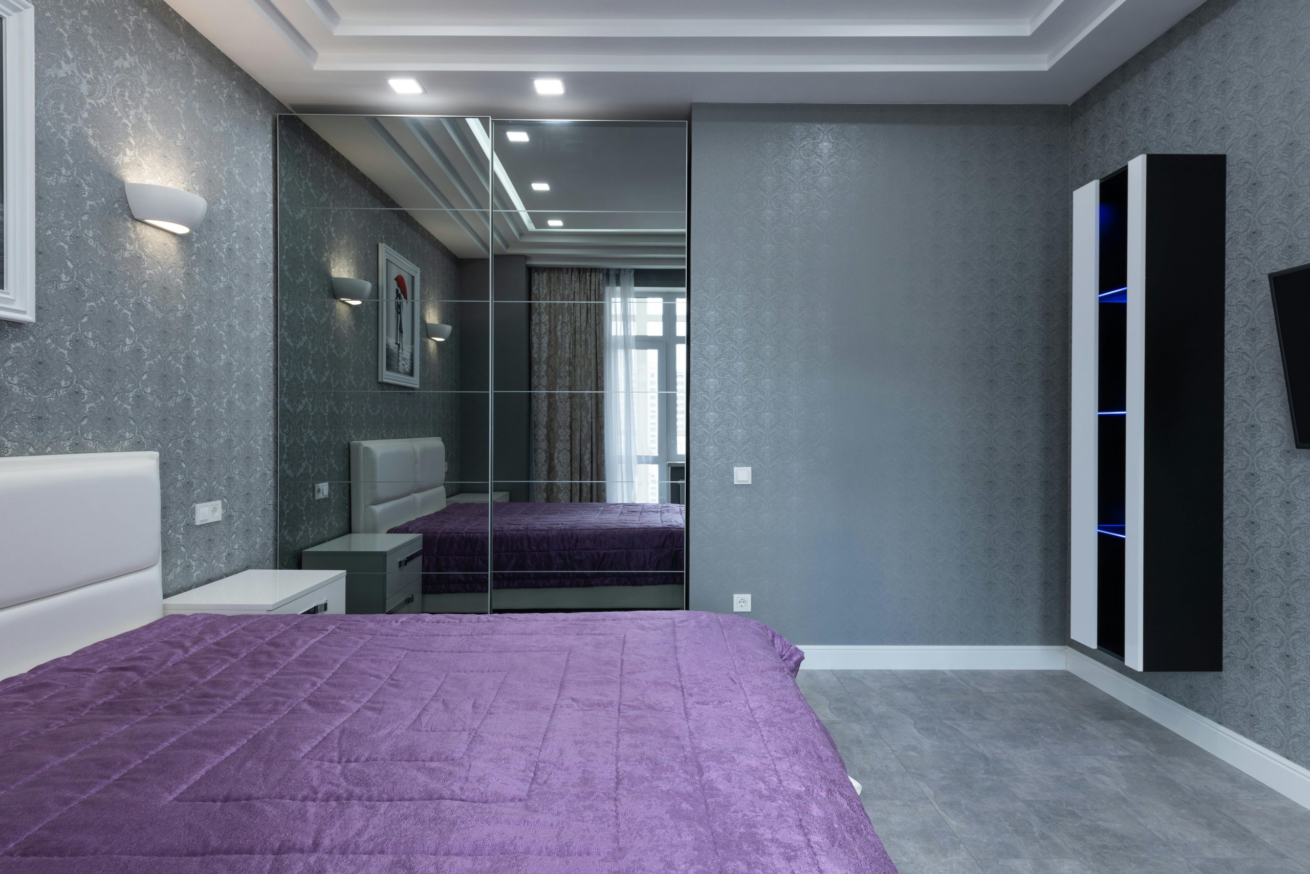 Comfortable bed with purple coverlet placed near wardrobe with mirrors in modern bedroom in flat