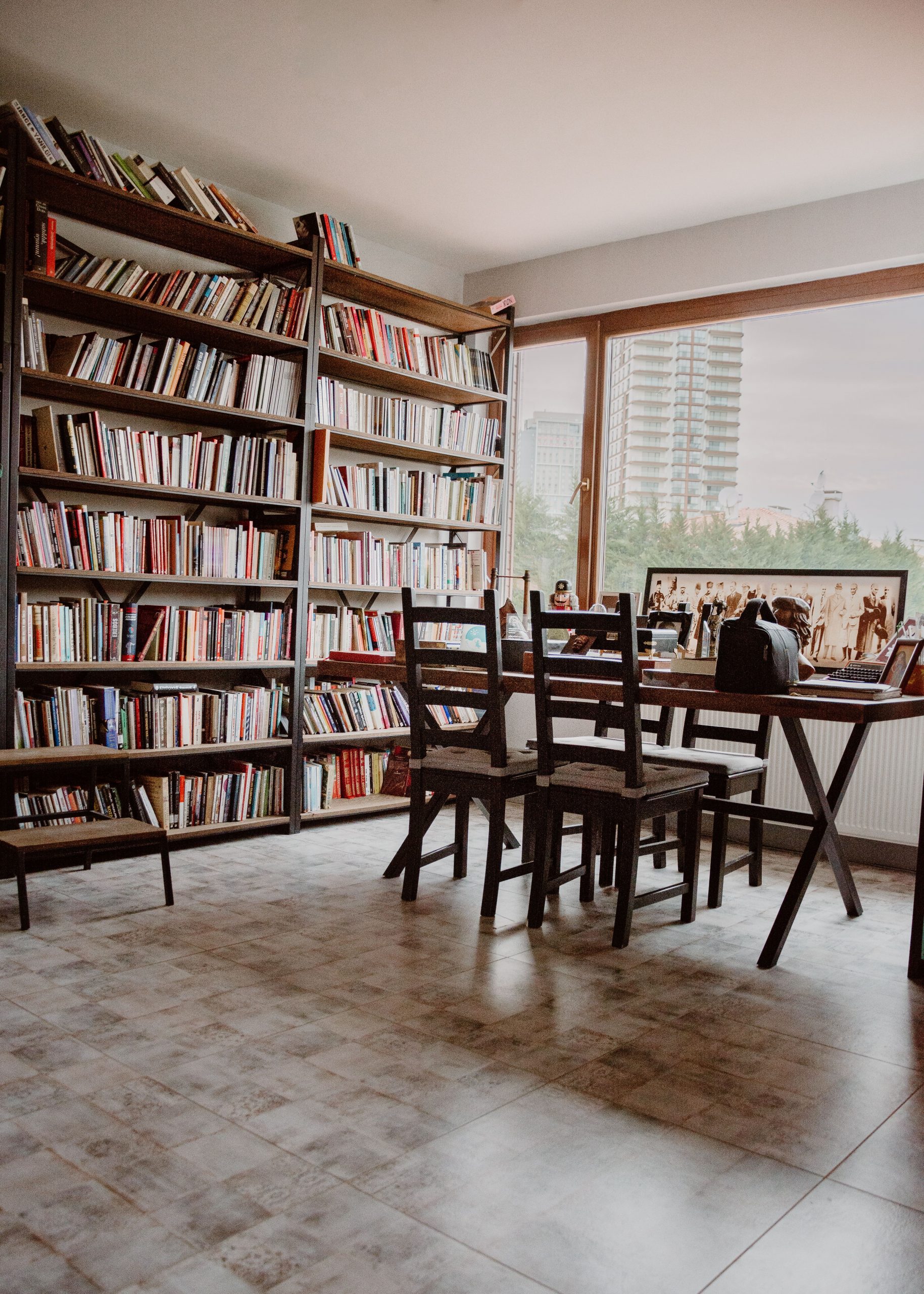 Cozy modern library with bookshelves, wooden chairs, and a city view through large windows.