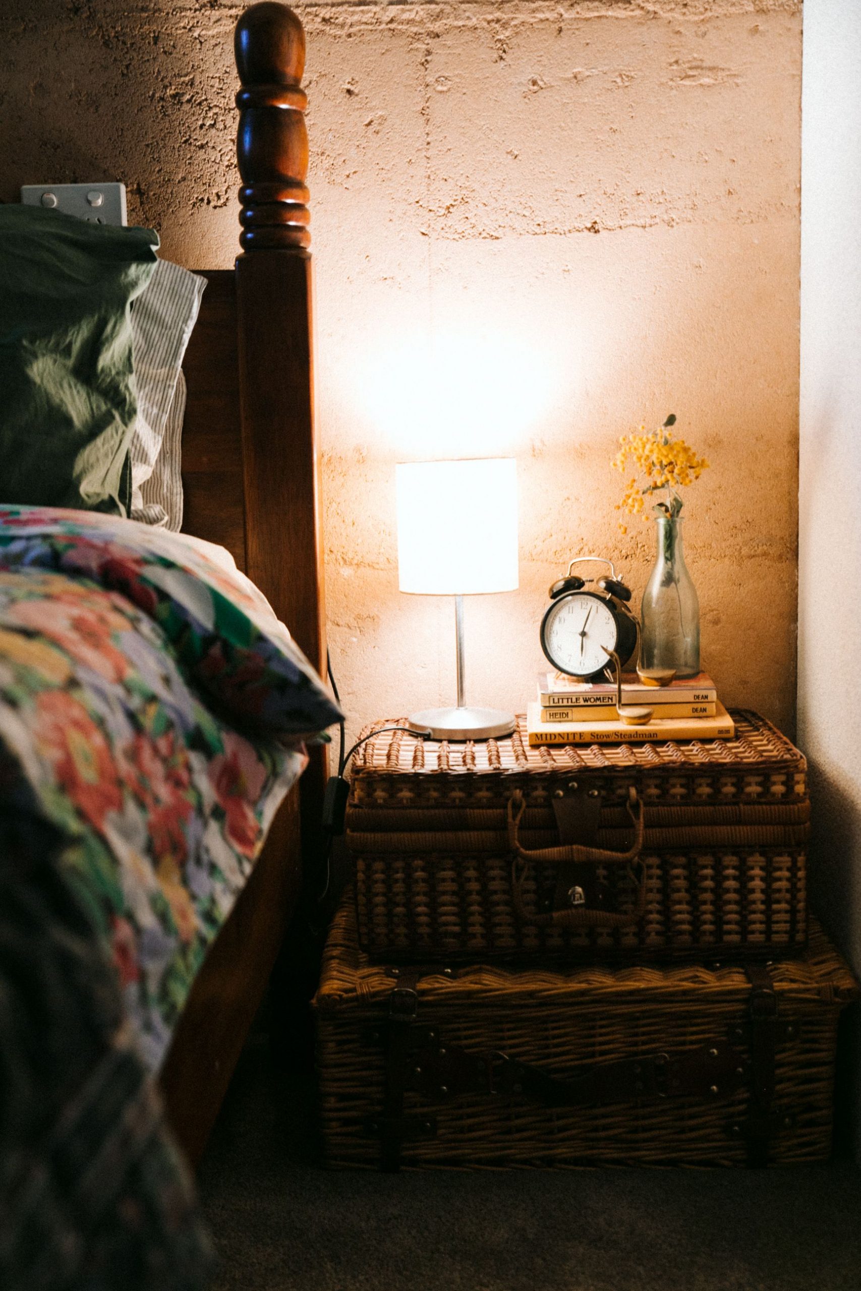 Domestic cozy bed near rustic bedside table made of wicker suitcases with glowing lamp in evening