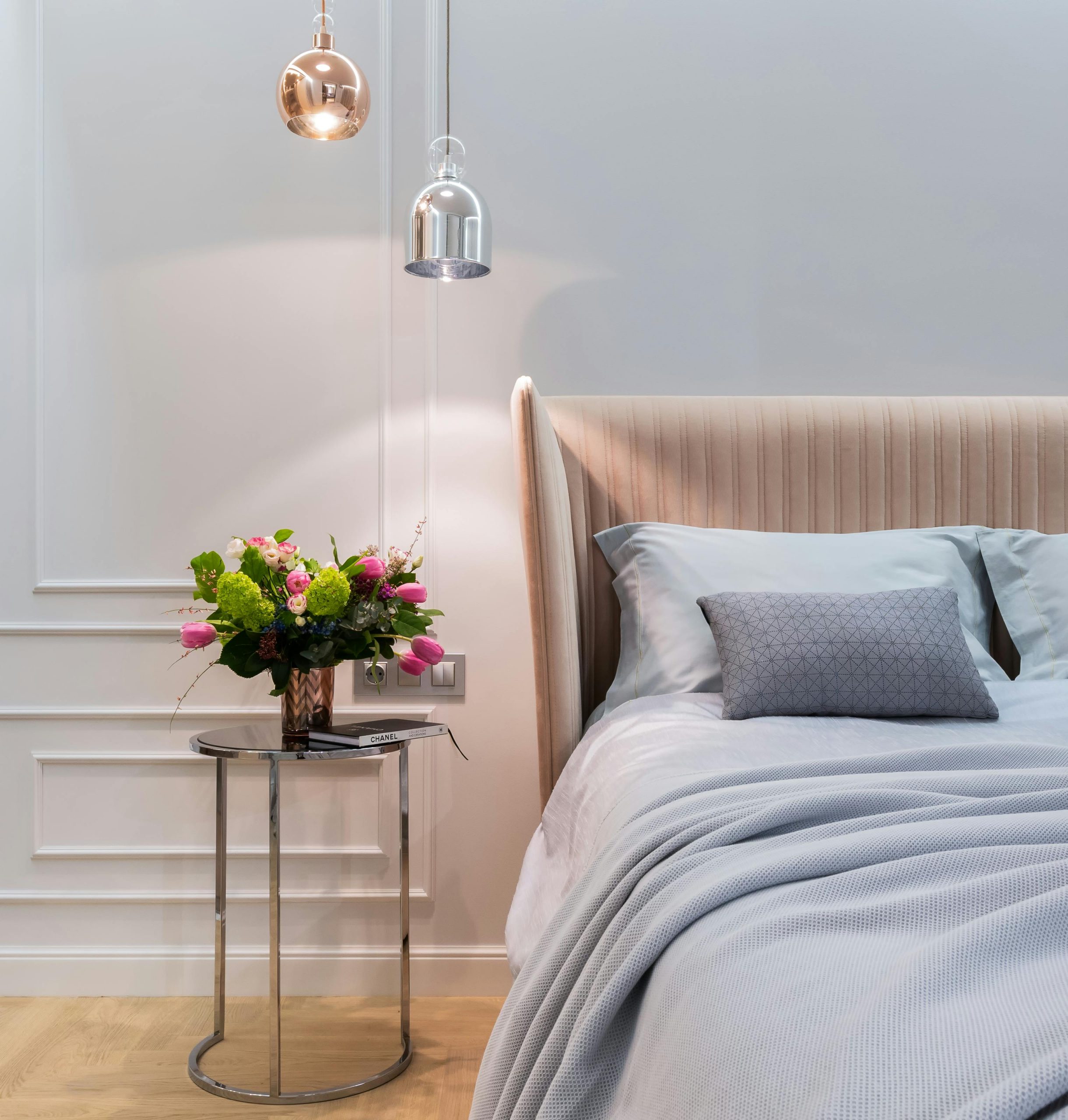Interior of modern light bedroom with pillows and blanket on bed near round table with bouquet of flowers in vase under lamps