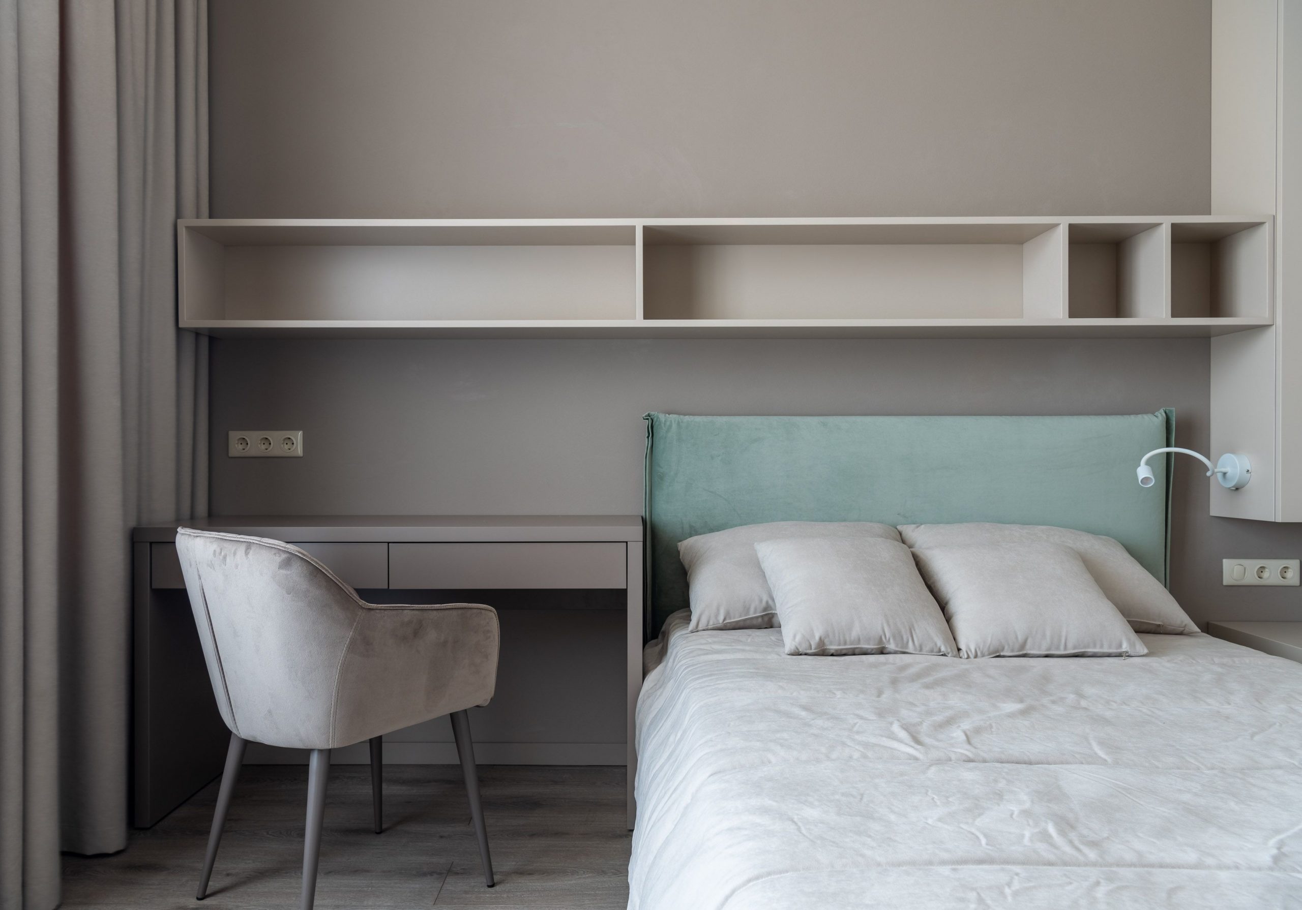 Interior of modern light bedroom with pillows on bed near table with chair under shelves next to curtains
