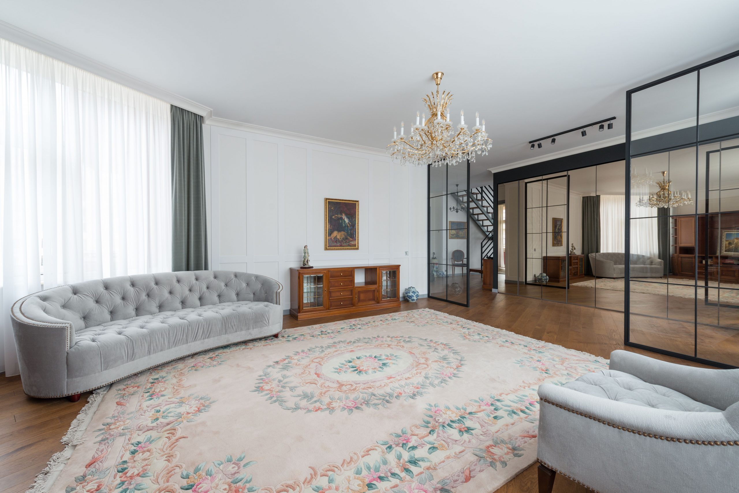 Interior of modern spacious living room with comfortable gray sofa and armchair and carpet with glass walls and big mirror