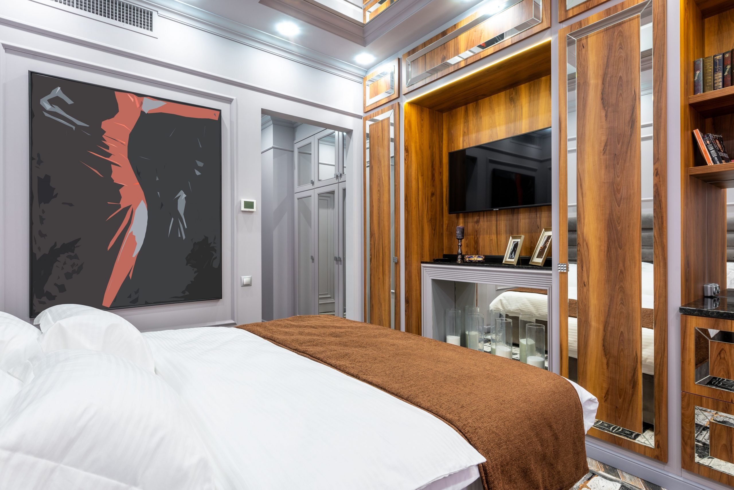 Modern bedroom interior with bed between painting and books on shelves illuminated by lights