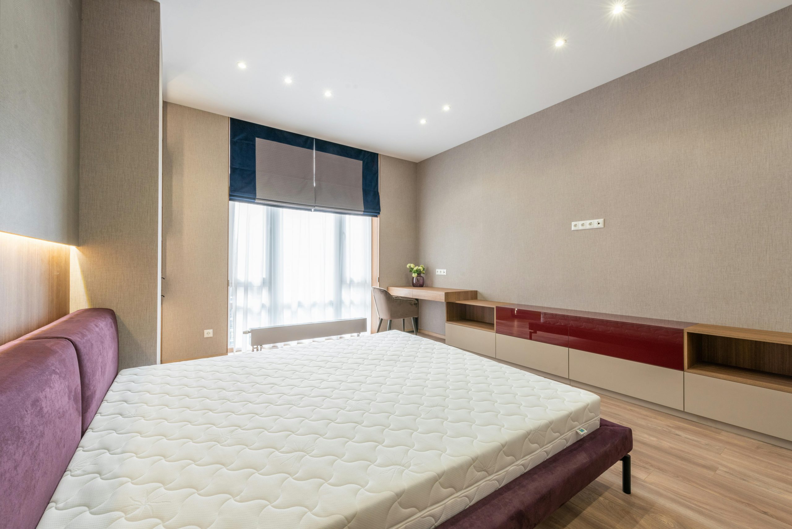 Modern bedroom with white mattress on bed placed against wooden cabinet and table with flowerpot at wall with roller blind on window