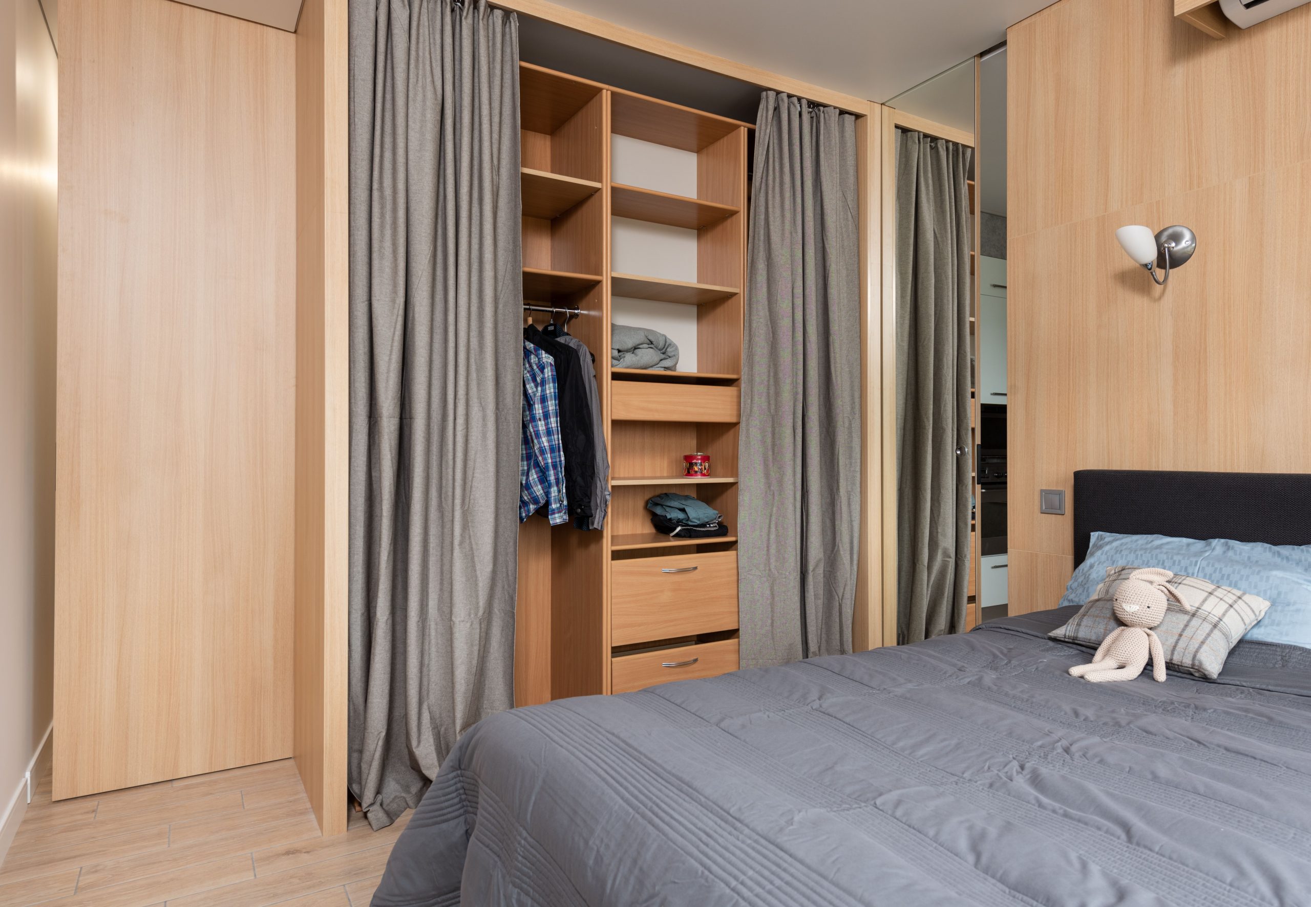 Modern room interior with bed with pillows and soft rabbit toy near wooden wardrobe with curtains and shelves near clothes on hangers in bright home