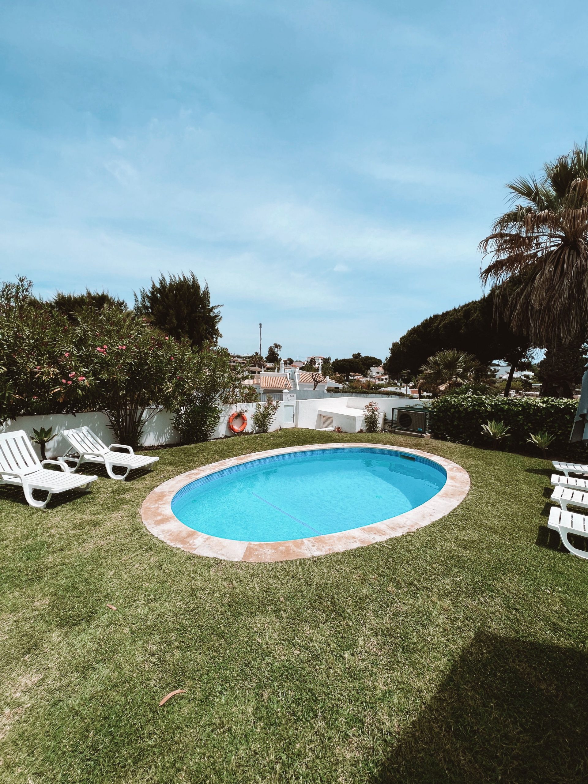 Serene outdoor scene featuring an oval pool surrounded by lush greenery and lounge chairs under a blue sky.