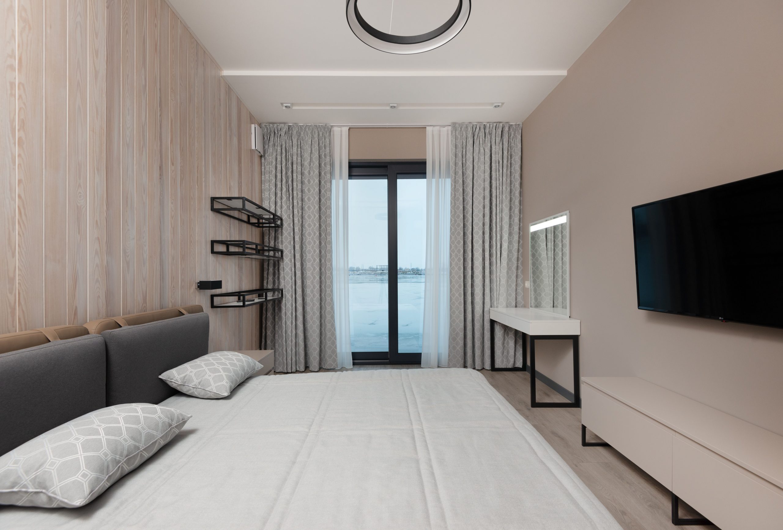 Soft bed with cushions and modern TV hanging on wall over cabinet in minimalist bedroom with vanity table and curtains