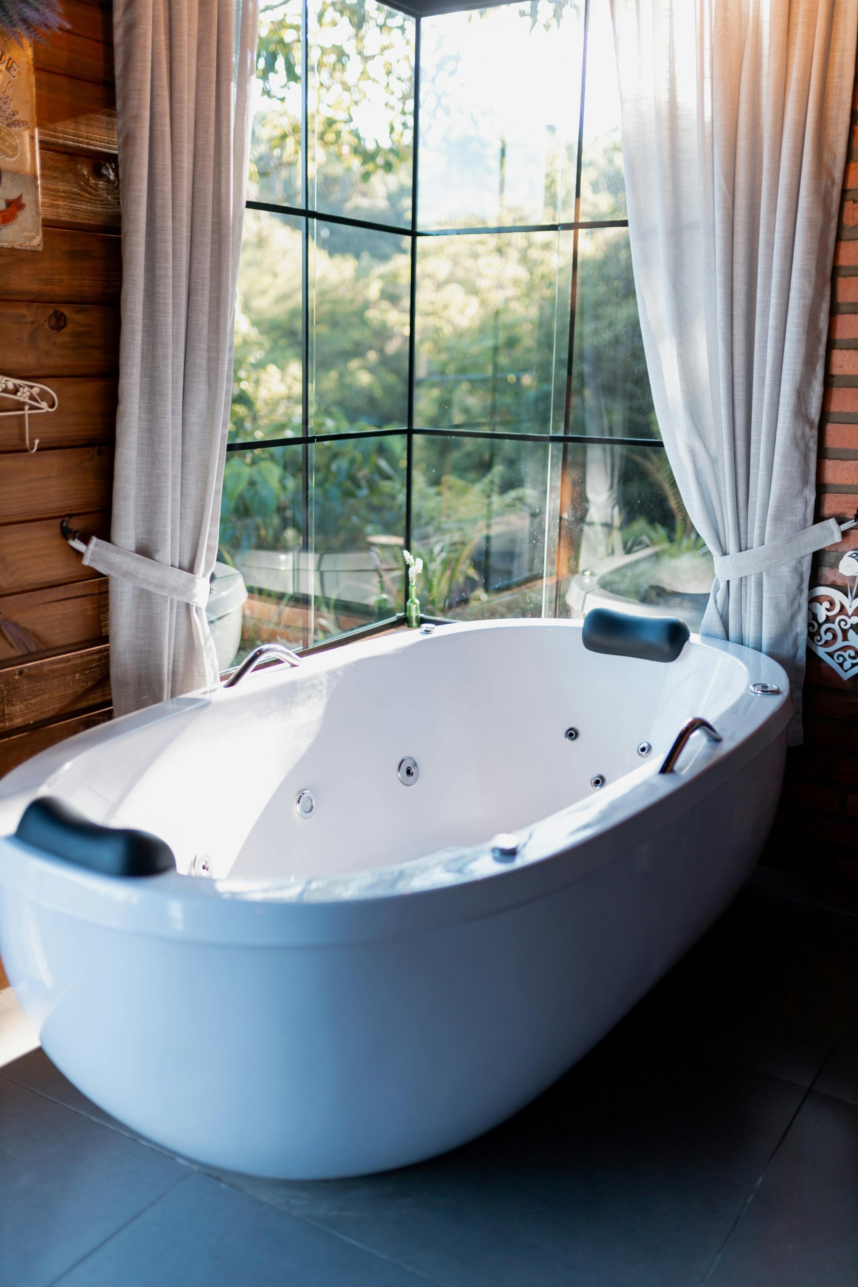 Spacious white bathtub in a sunlit bathroom with large windows and curtains, offering a serene escape.