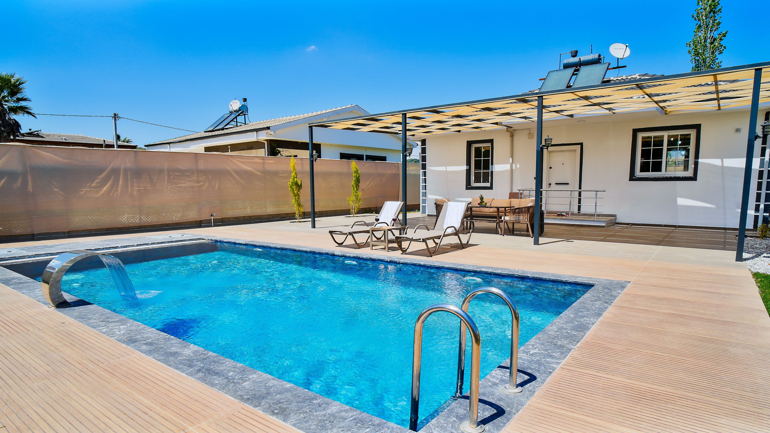 Sunny day view of a luxurious outdoor pool and patio with modern lounge chairs and a pergola.