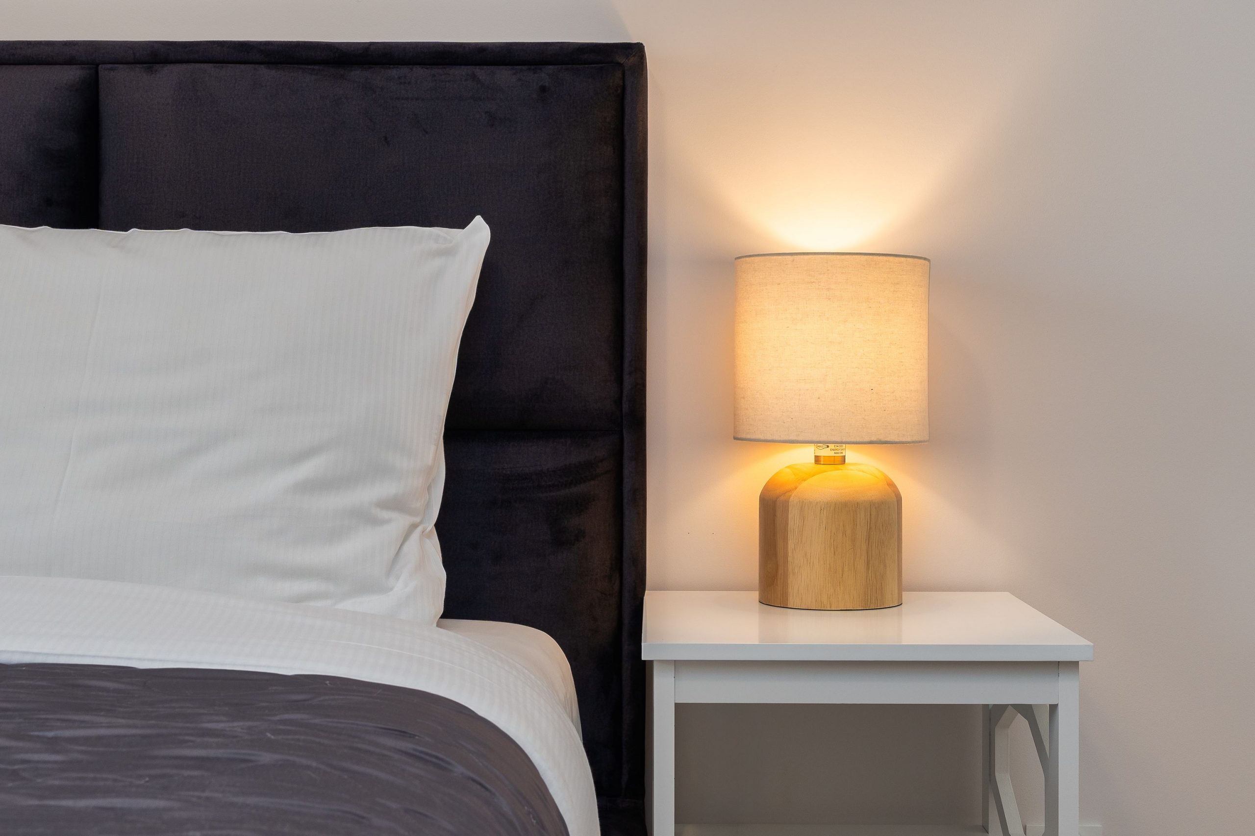 Warmly lit bedroom lamp with modern design on a white nightstand beside a comfortable bed.