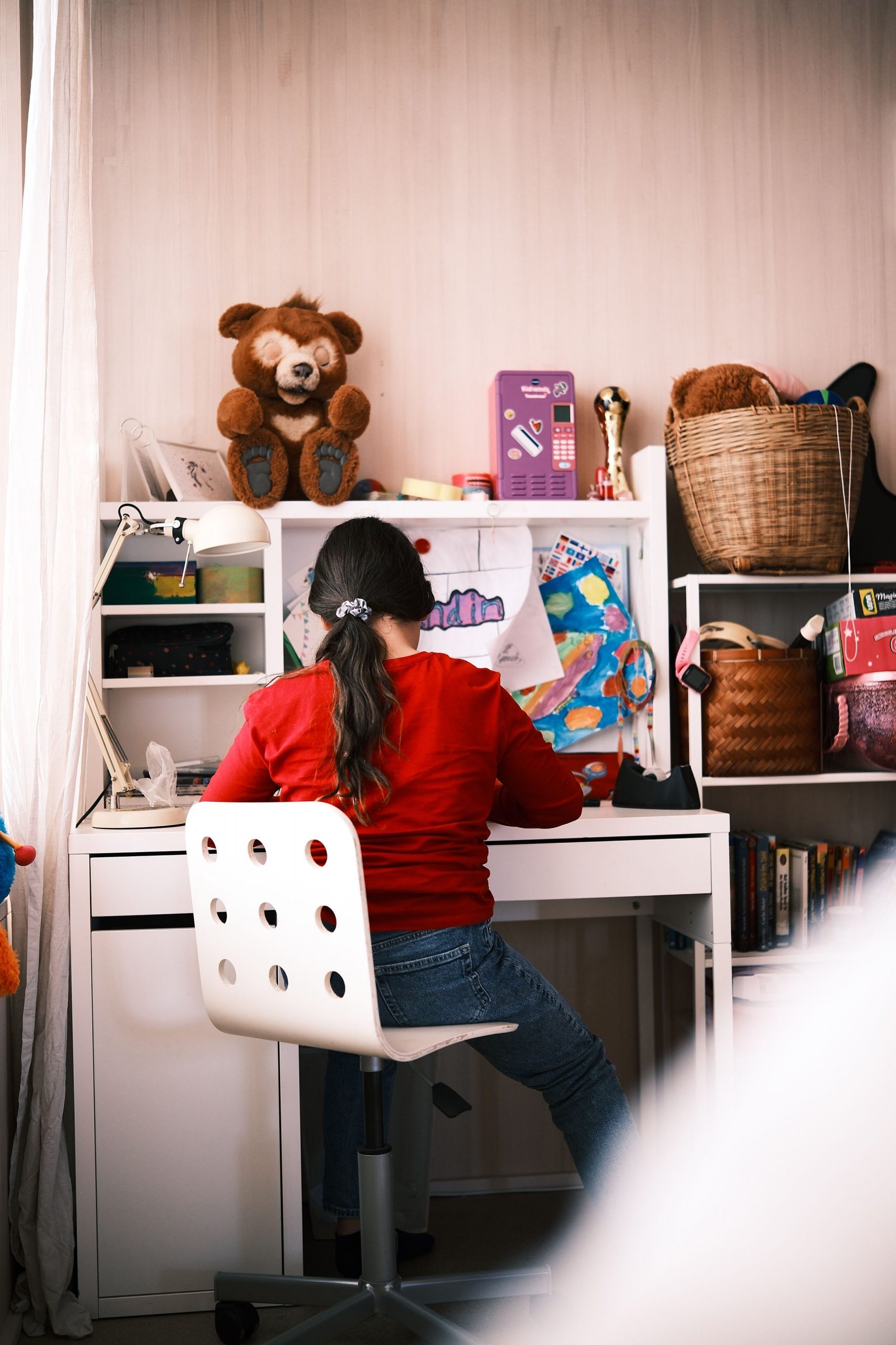 Young girl studying at a desk in a cozy bedroom, surrounded by toys and books.
