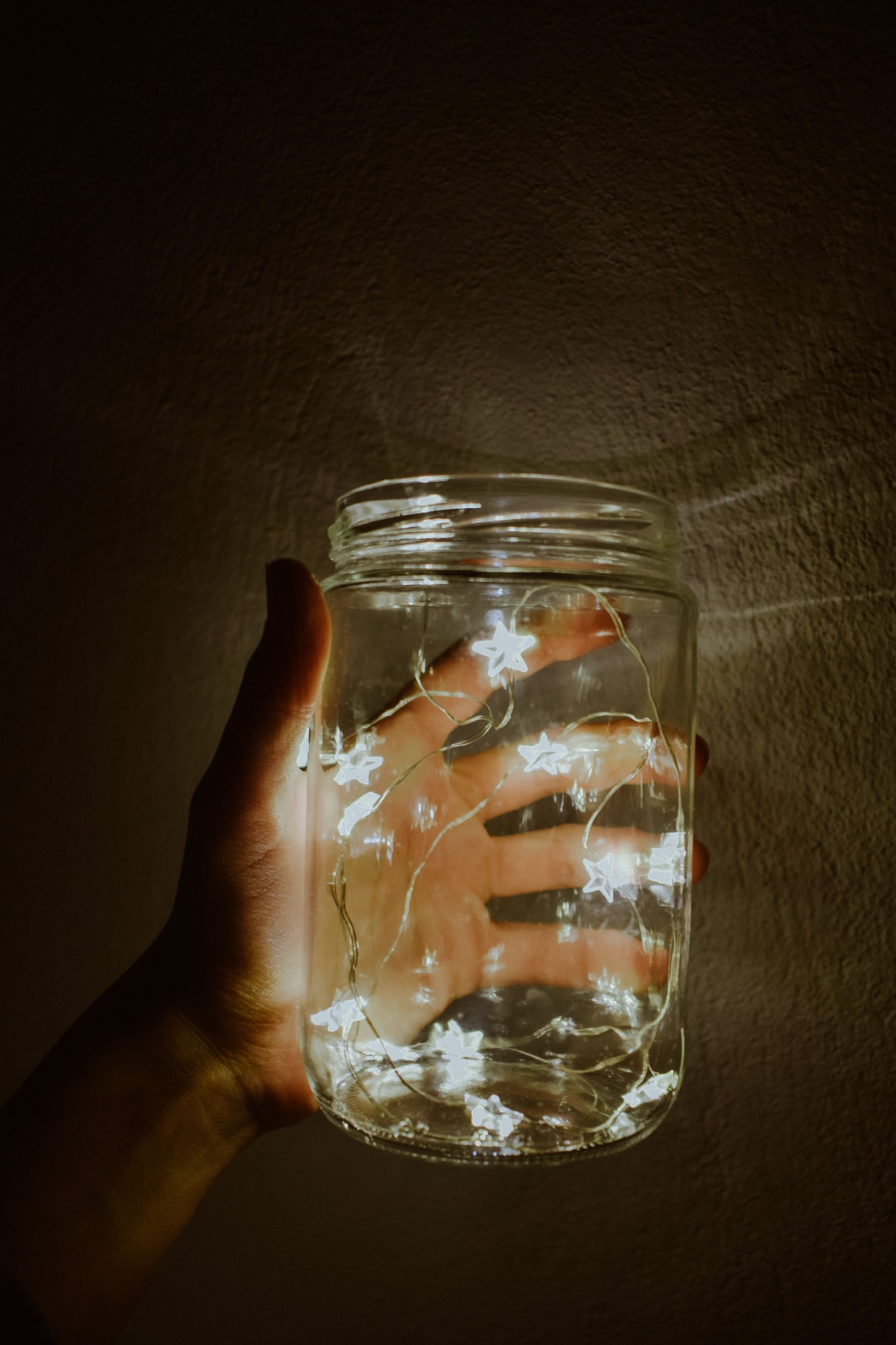A hand holding a mason jar filled with glowing star-shaped fairy lights against a dark background.