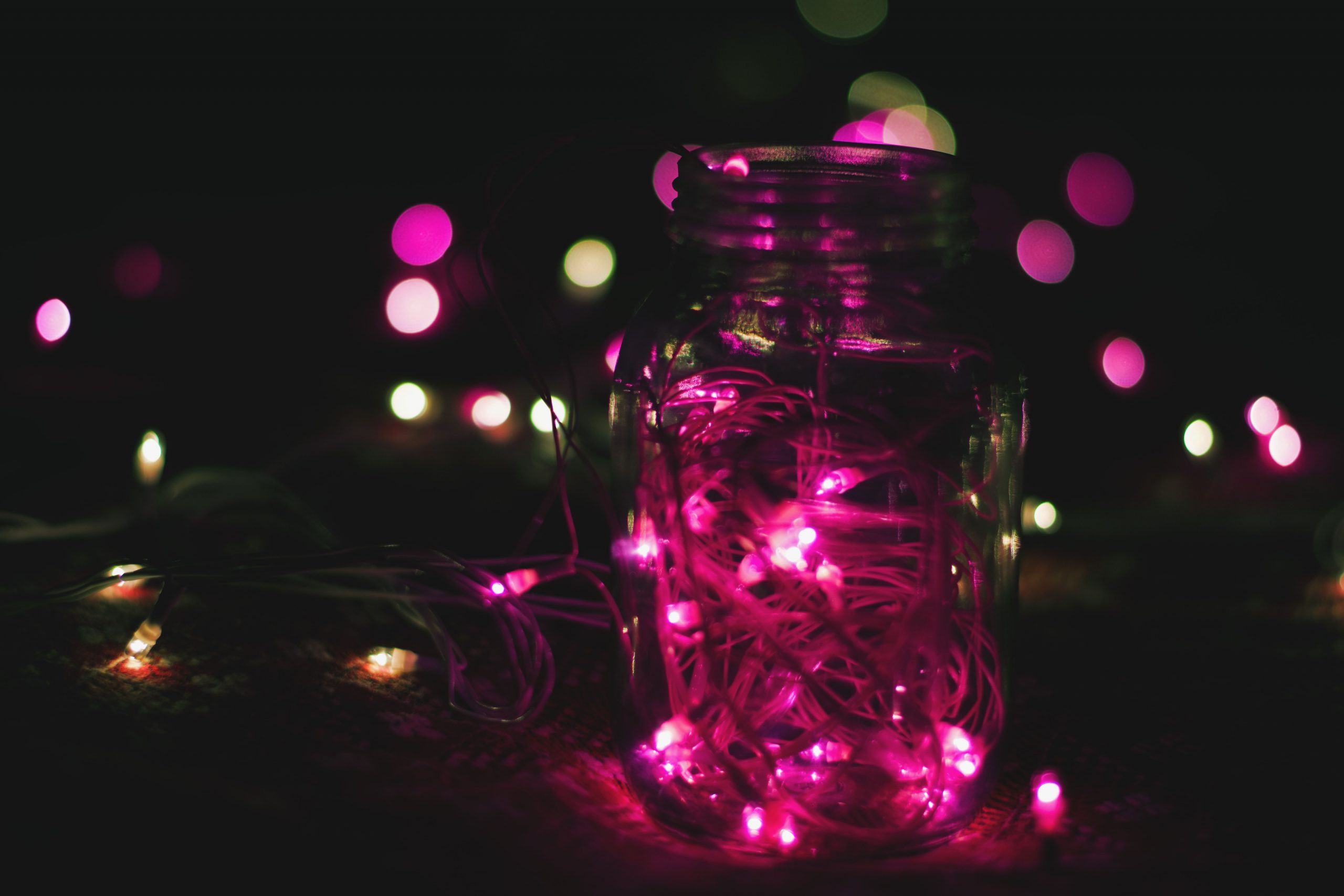 A mason jar filled with glowing pink neon string lights surrounded by bokeh in a dark setting.