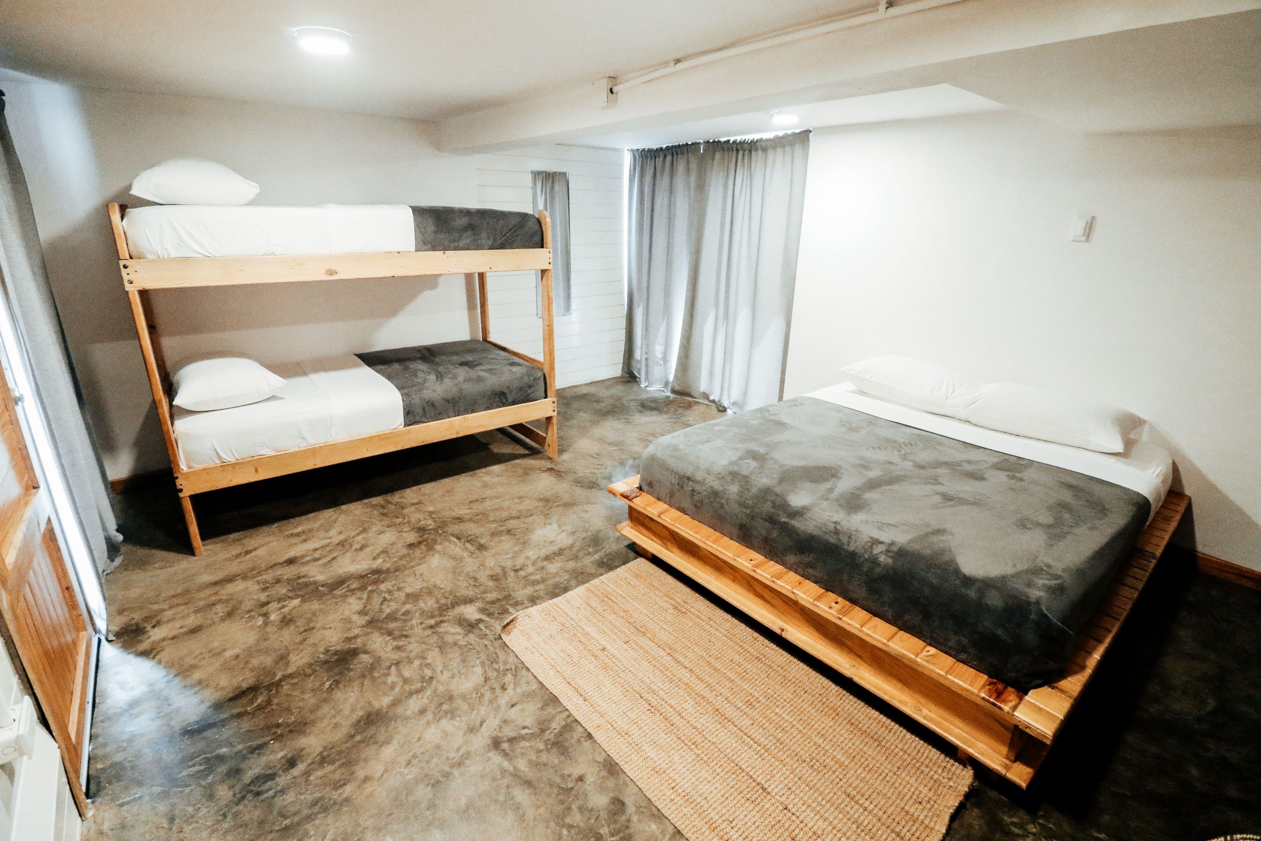 A minimalistic bedroom featuring a wooden bunk bed and platform bed on a concrete floor.