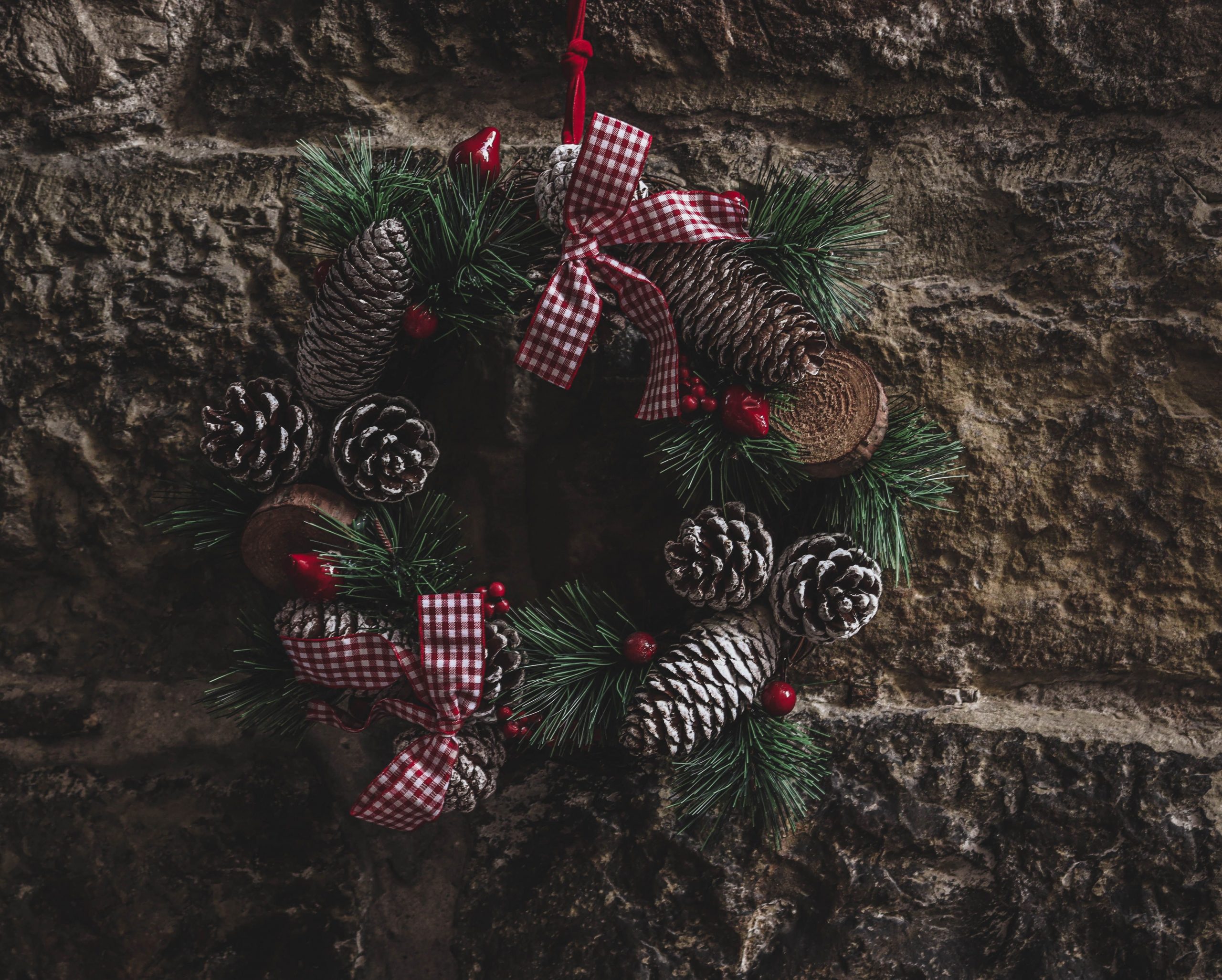 A rustic Christmas wreath with pinecones and red ribbons hangs on a stone wall.