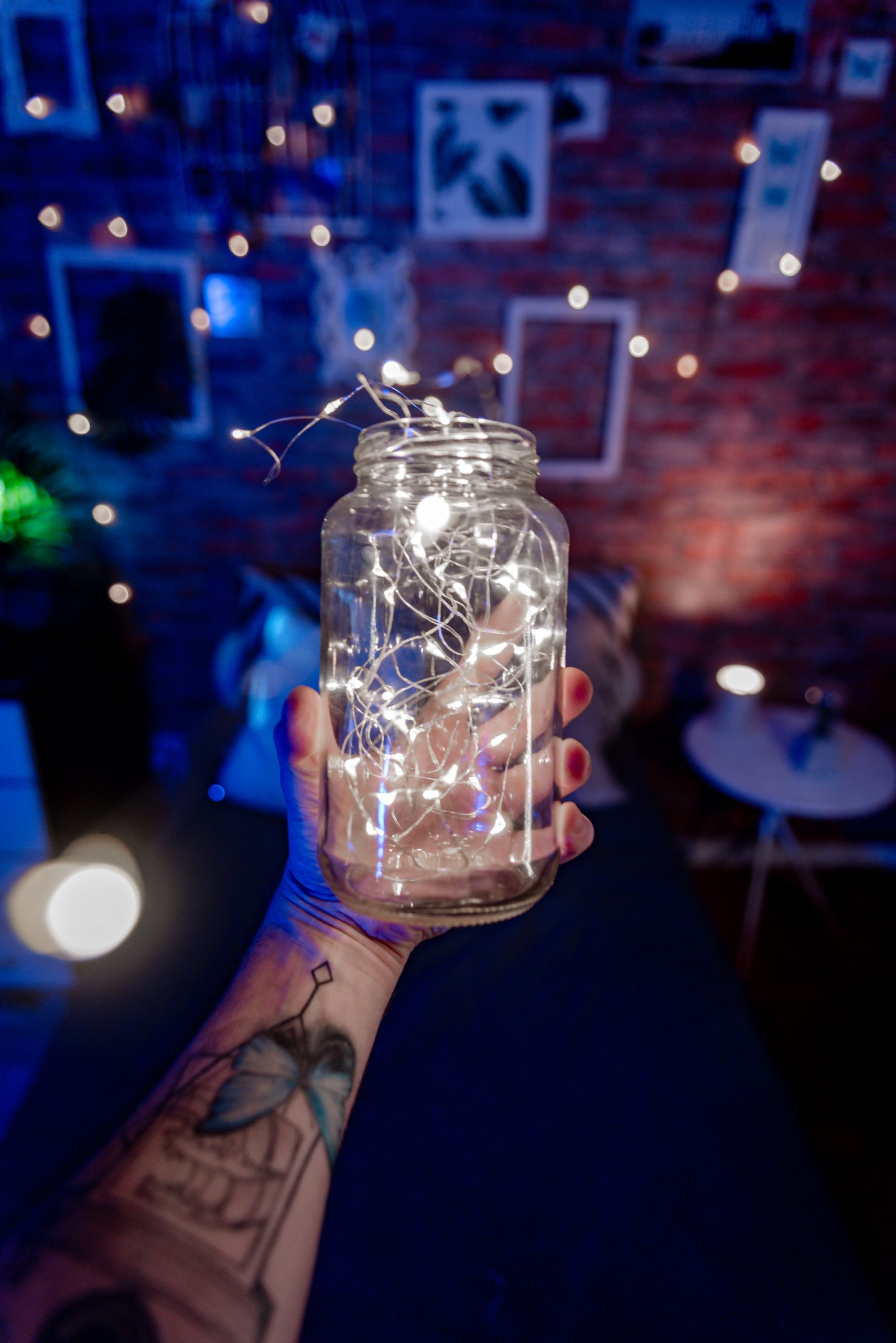 A tattooed hand holds a glowing jar of string lights in a cozy room with warm decoration.