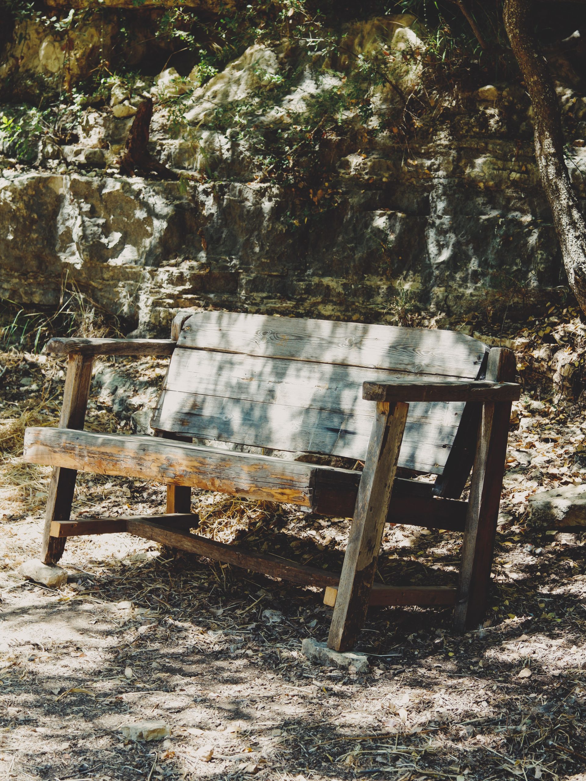 An old wooden bench under dappled sunlight, set against a rocky background outdoors.