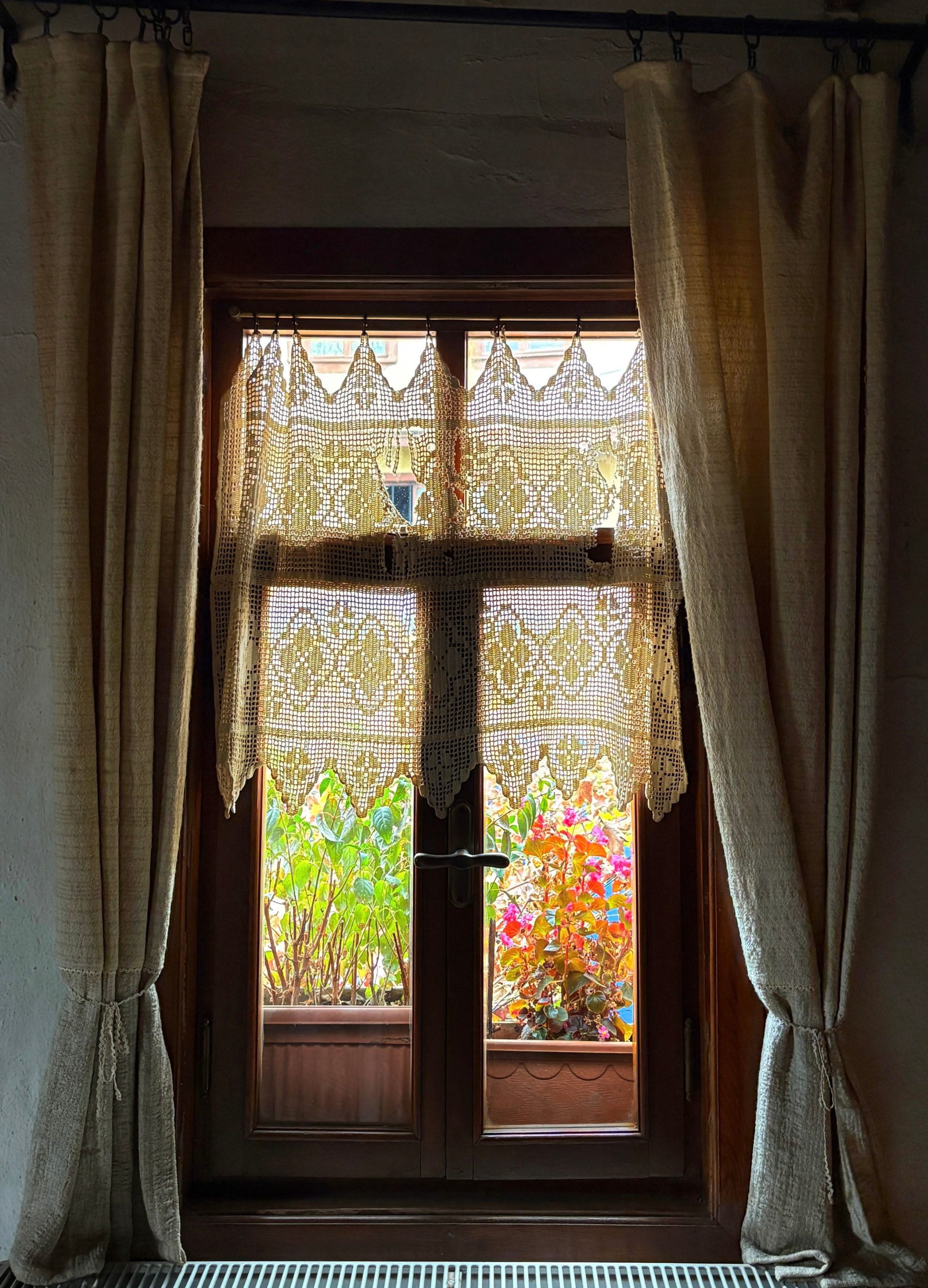 Charming view through a wooden window with lace curtains and colorful flowers outside.