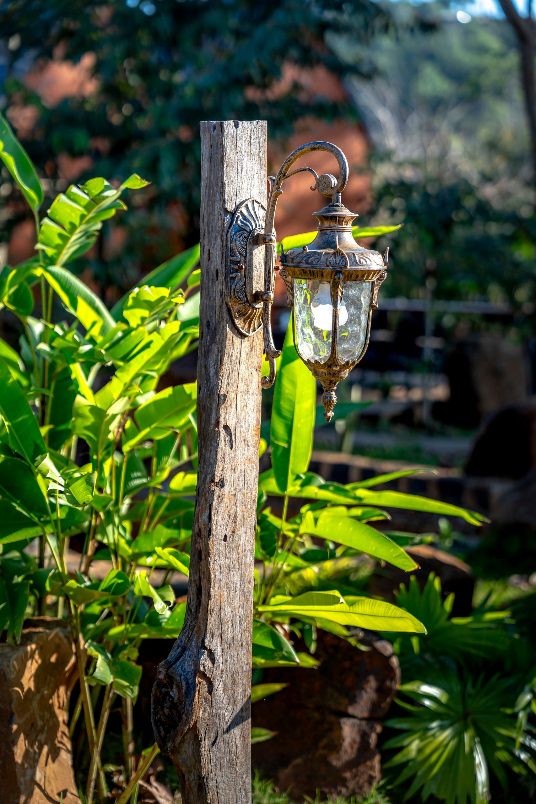 Close-up of an antique lamp hanging on a weathered wooden pole in a lush garden setting.