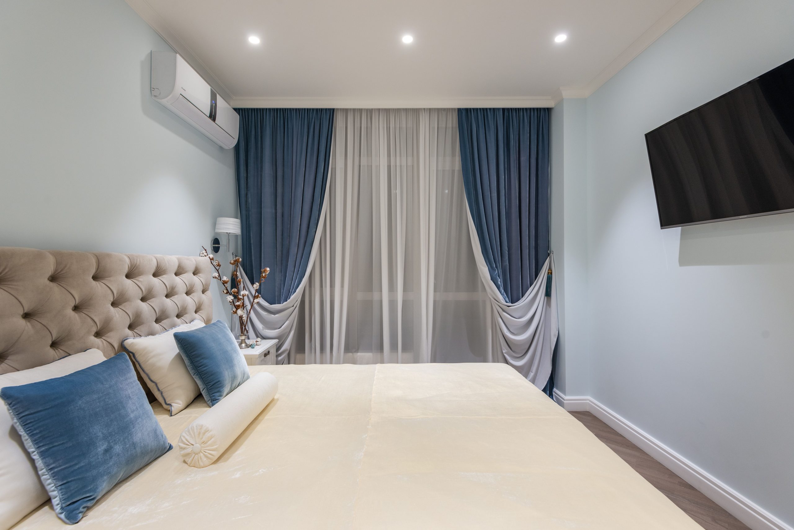 Comfortable bed with soft headboard and modern TV hanging on wall in classic styled bedroom decorated with blue velour curtains and cushions
