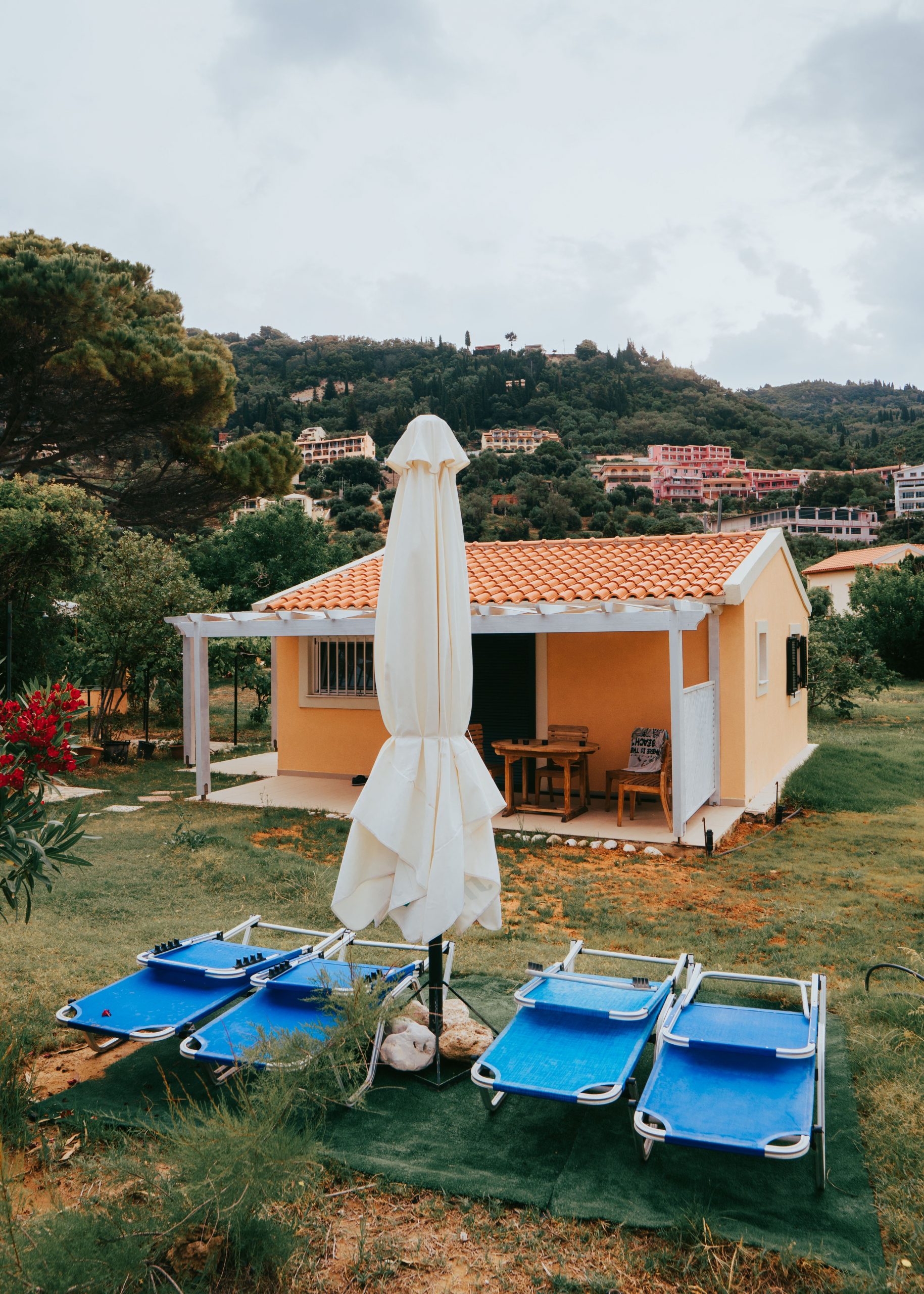 Cozy village cottage with sun loungers and umbrella amidst lush greenery.