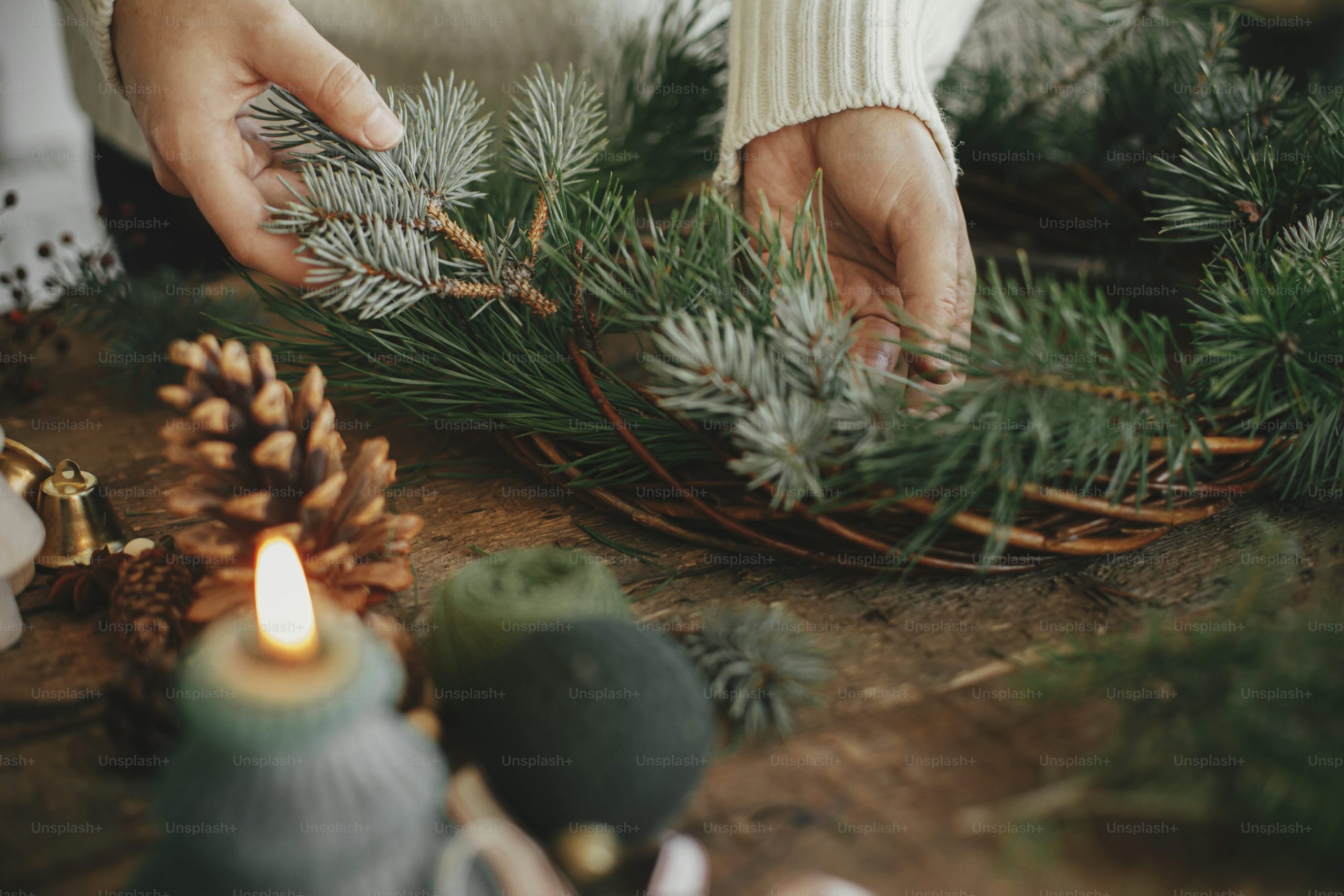 Making rustic christmas wreath close up. Woman hands holding fir branch and arranging christmas wreath on rustic wooden background with candle, pine cones, thread. Winter holiday preparations photo